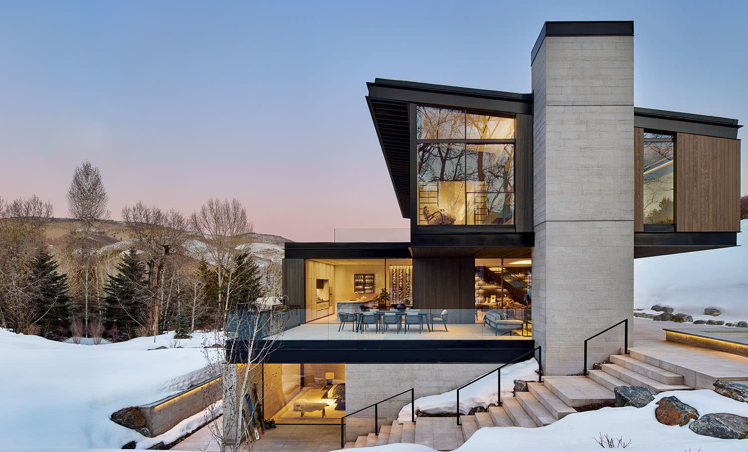 Modern house in snowy landscape at sunset, featuring large glass windows and a balcony. Warm lights inside contrast with the cool, serene outdoor setting.