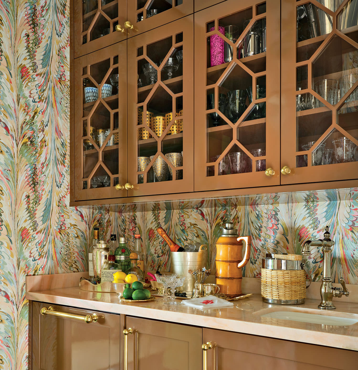 Warm, retro-themed kitchen with geometric glass cabinets, patterned wallpaper, and a counter adorned with drinks, lemons, and elegant glassware.