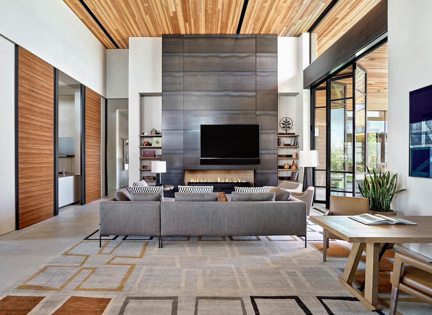 Modern living room with high wooden ceilings and large glass doors. Gray sofas face a wall-mounted TV above a sleek fireplace. Light, airy ambiance.