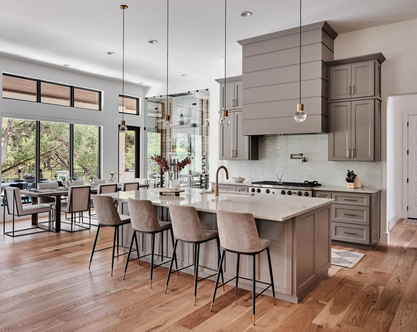 Spacious modern kitchen with gray cabinets, a large island with beige stools, wood floors, pendant lights, and a view of trees through large windows.