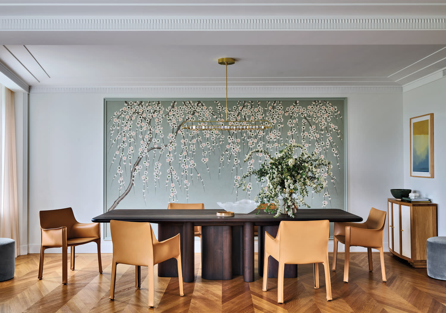 Elegant dining room with a dark wooden table, tan chairs, and a floral feature wall. A chandelier hangs above. The space feels modern and serene.