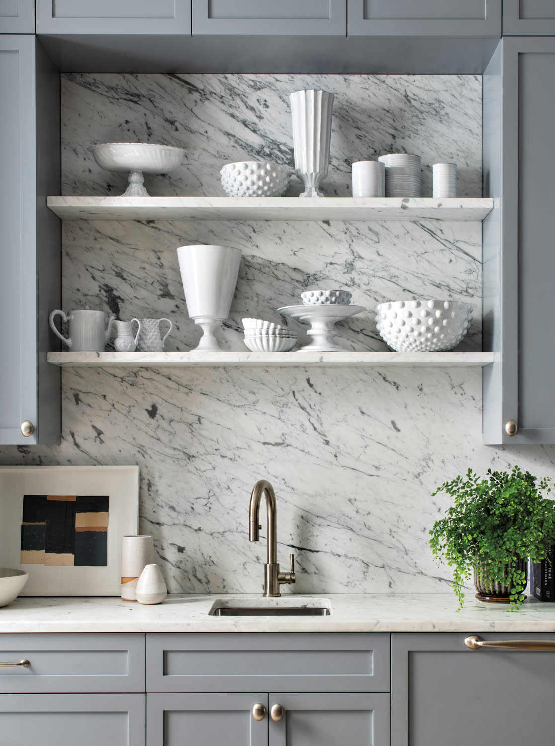 Stylish kitchen with grey cabinets, marble backsplash, open shelves with white ceramics, and a sleek bronze faucet. A potted plant adds greenery.