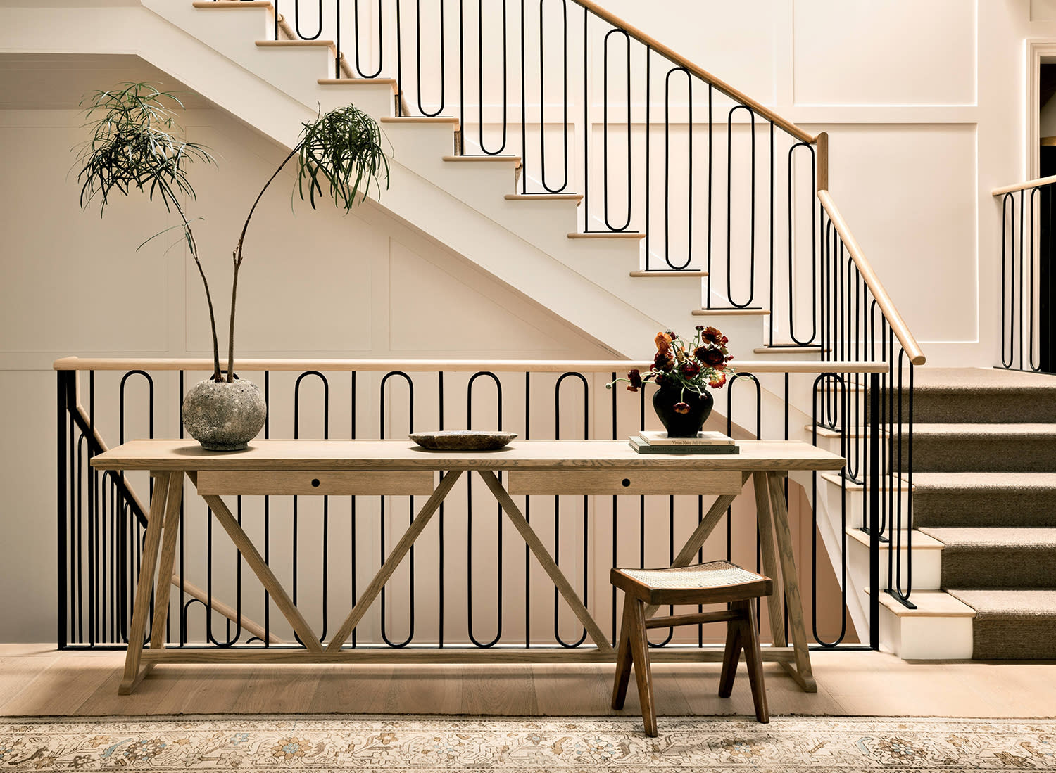 An entryway staircase with a wood table and matching stool set near a railing alongside a pale rug.