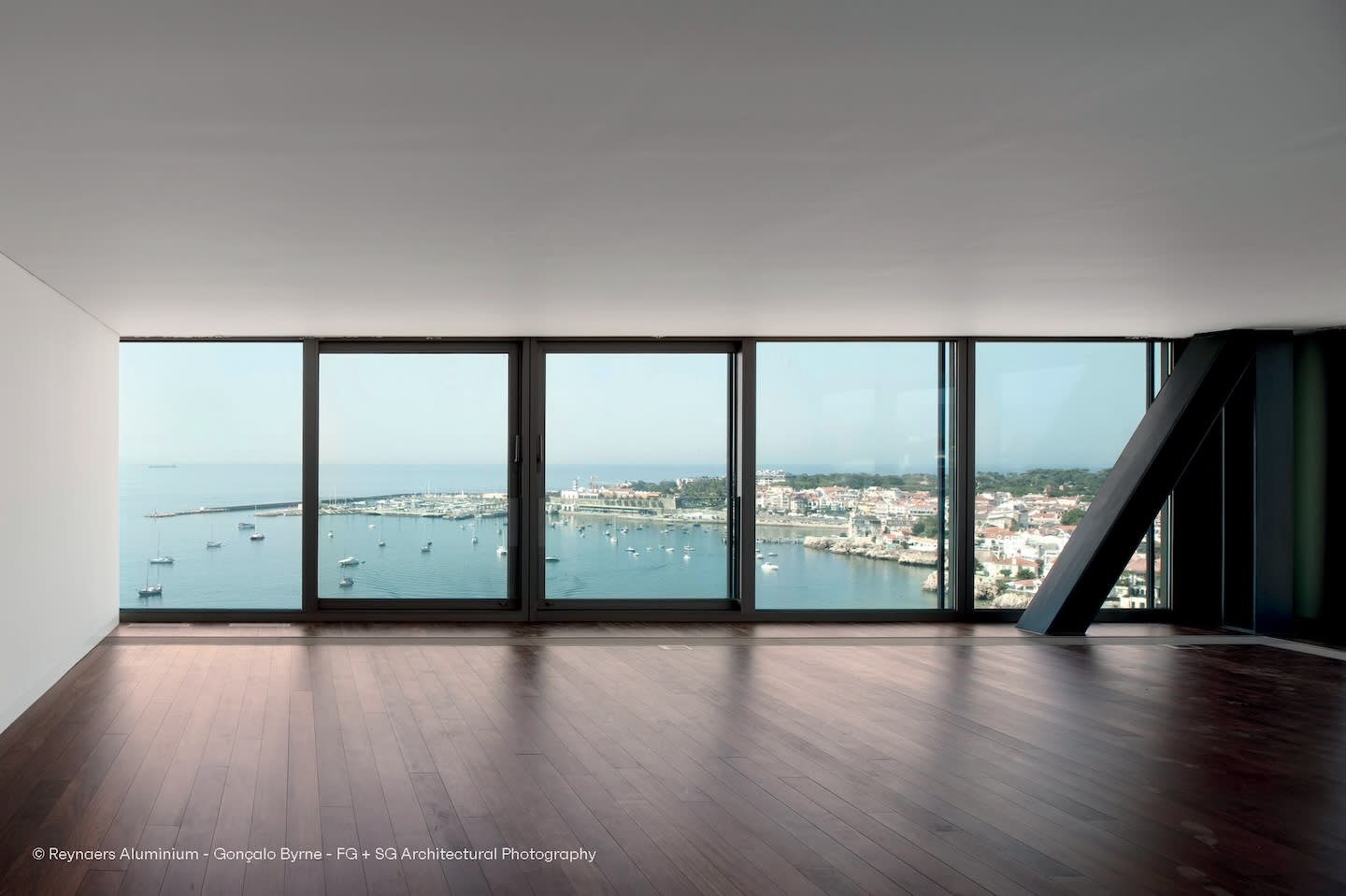 Spacious room with wooden floor, large floor-to-ceiling windows revealing a serene harbor view, boats, and distant buildings under a clear sky.