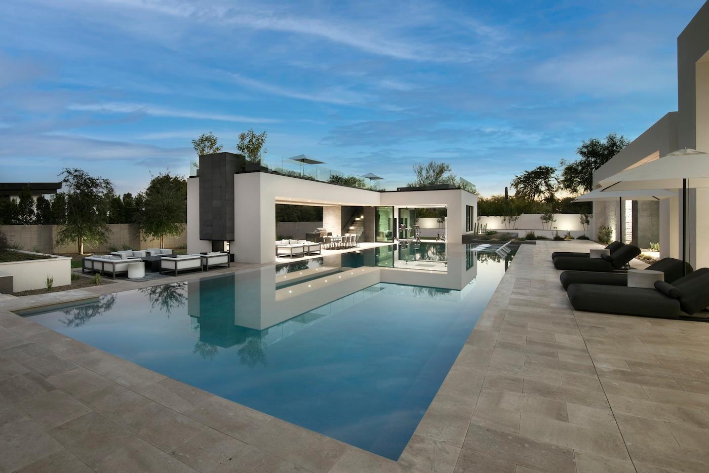 Luxurious modern poolside with sleek lounge chairs and a contemporary open-air structure. Evening sky reflects in the calm water, creating a serene ambiance.
