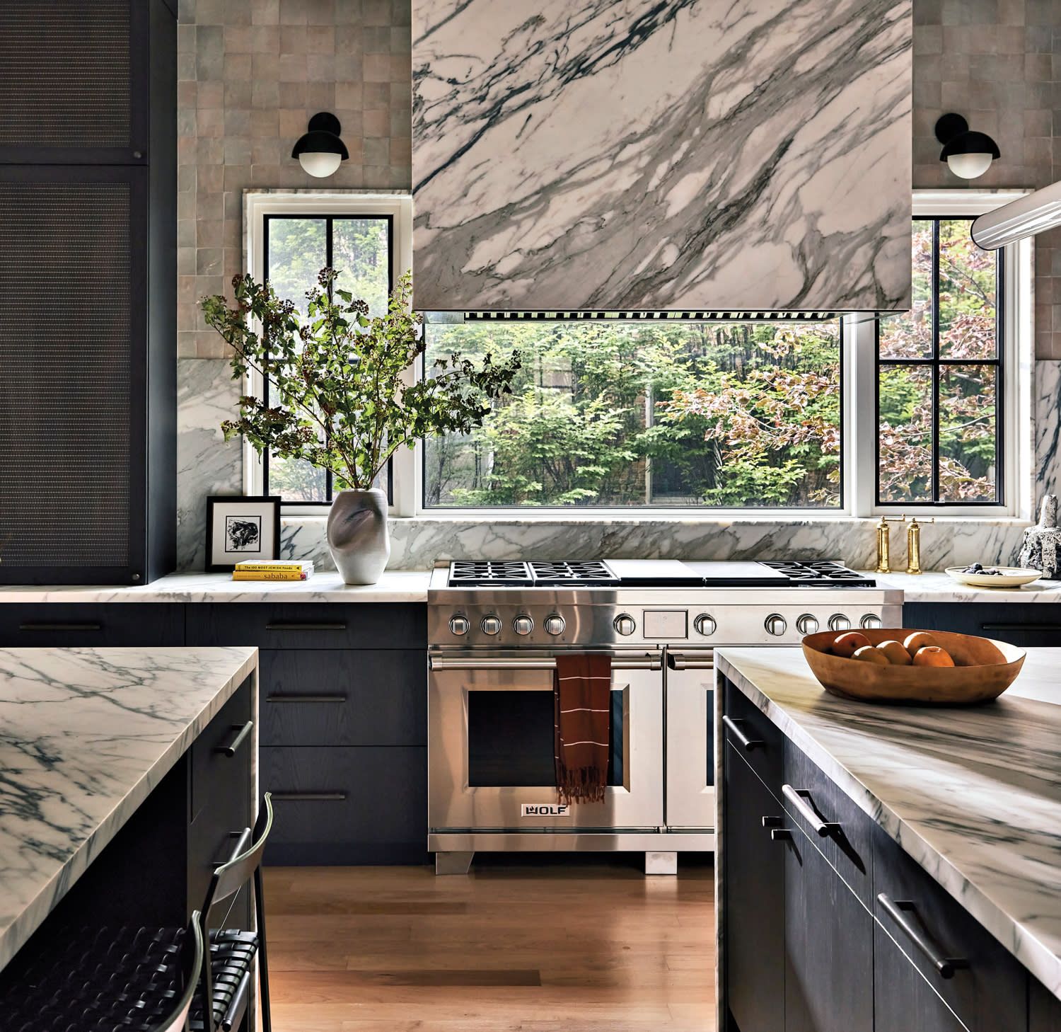 Elegant kitchen with marble countertops and backsplash, stainless steel oven, large window with greenery view, and a vase with branches on the counter.