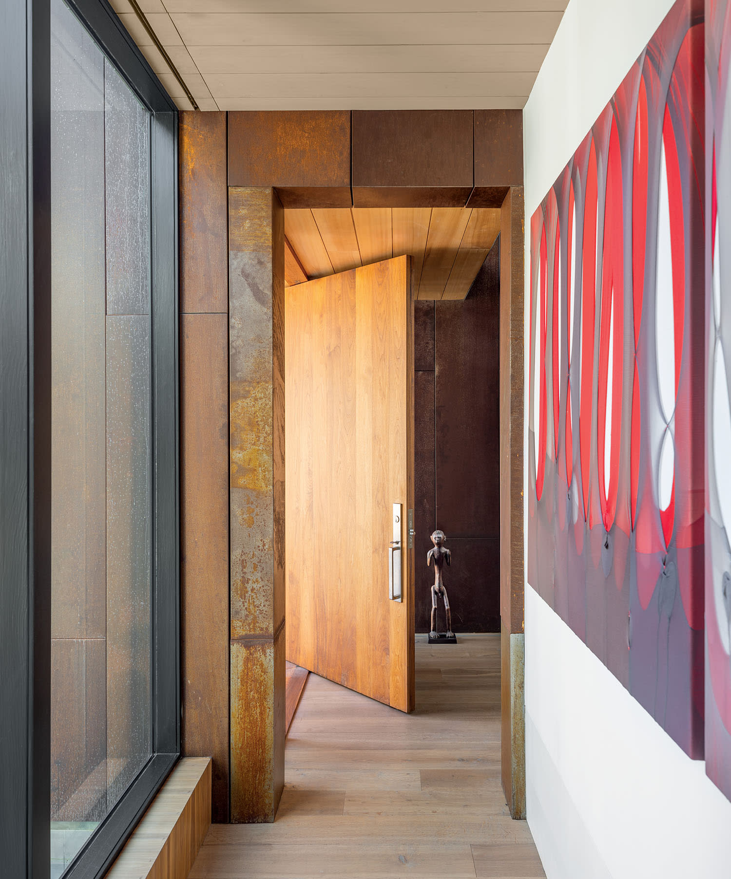 A modern hallway with a large wooden door slightly ajar, revealing a sculpture inside. The left wall features a large window, while red abstract art decorates the right.