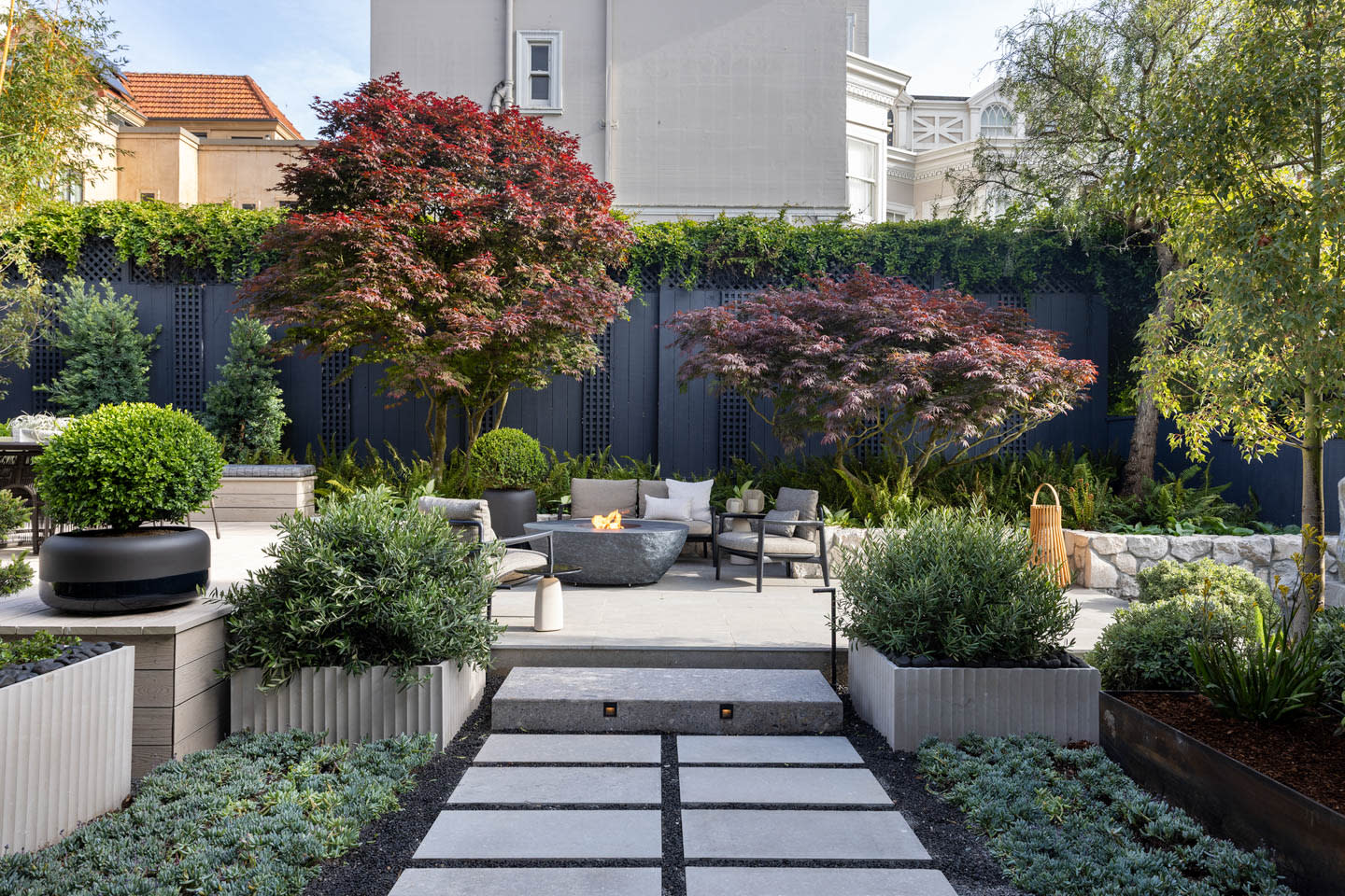 Modern garden patio with stone path, lush greenery, red maple trees, and cozy seating around a fire pit, exuding a tranquil, inviting atmosphere.