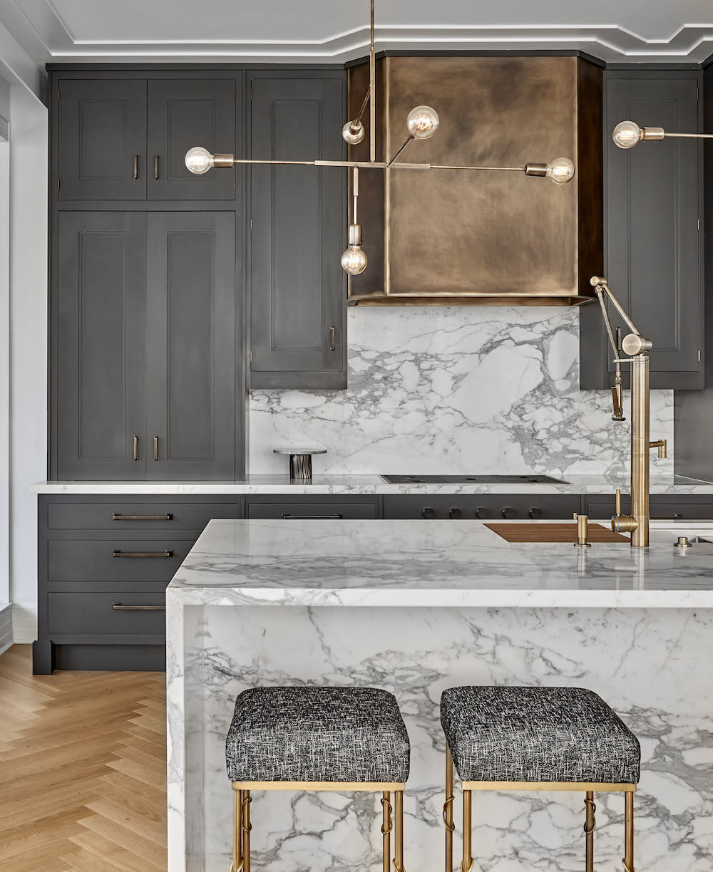 Dark hand painted cabinetry, brass hood and fixtures, sculptural pendant lighting hangs above the marble waterfall island.