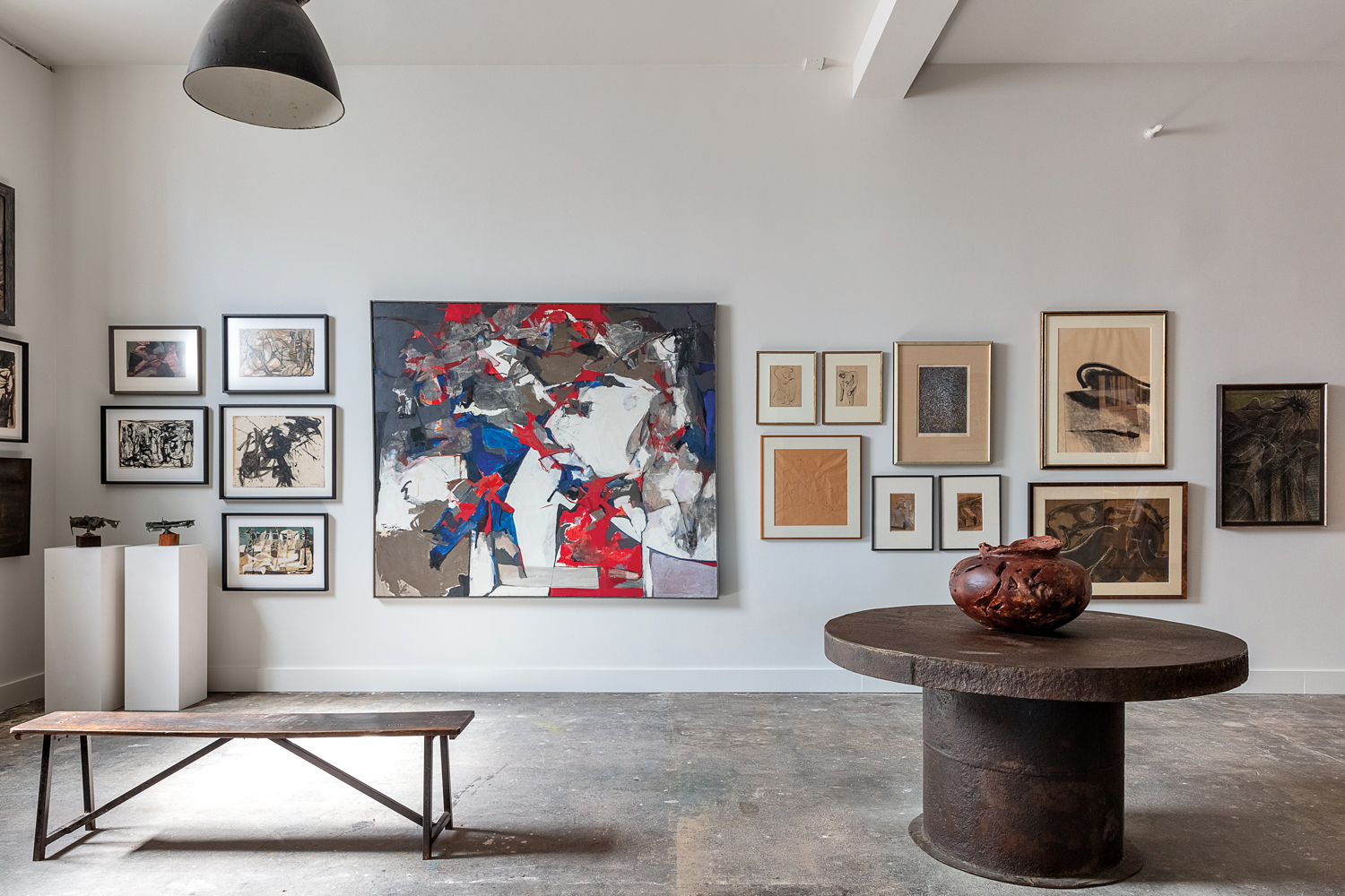 Minimalist gallery space with walls covered in artwork, a bench and rustic vessel atop a round table.