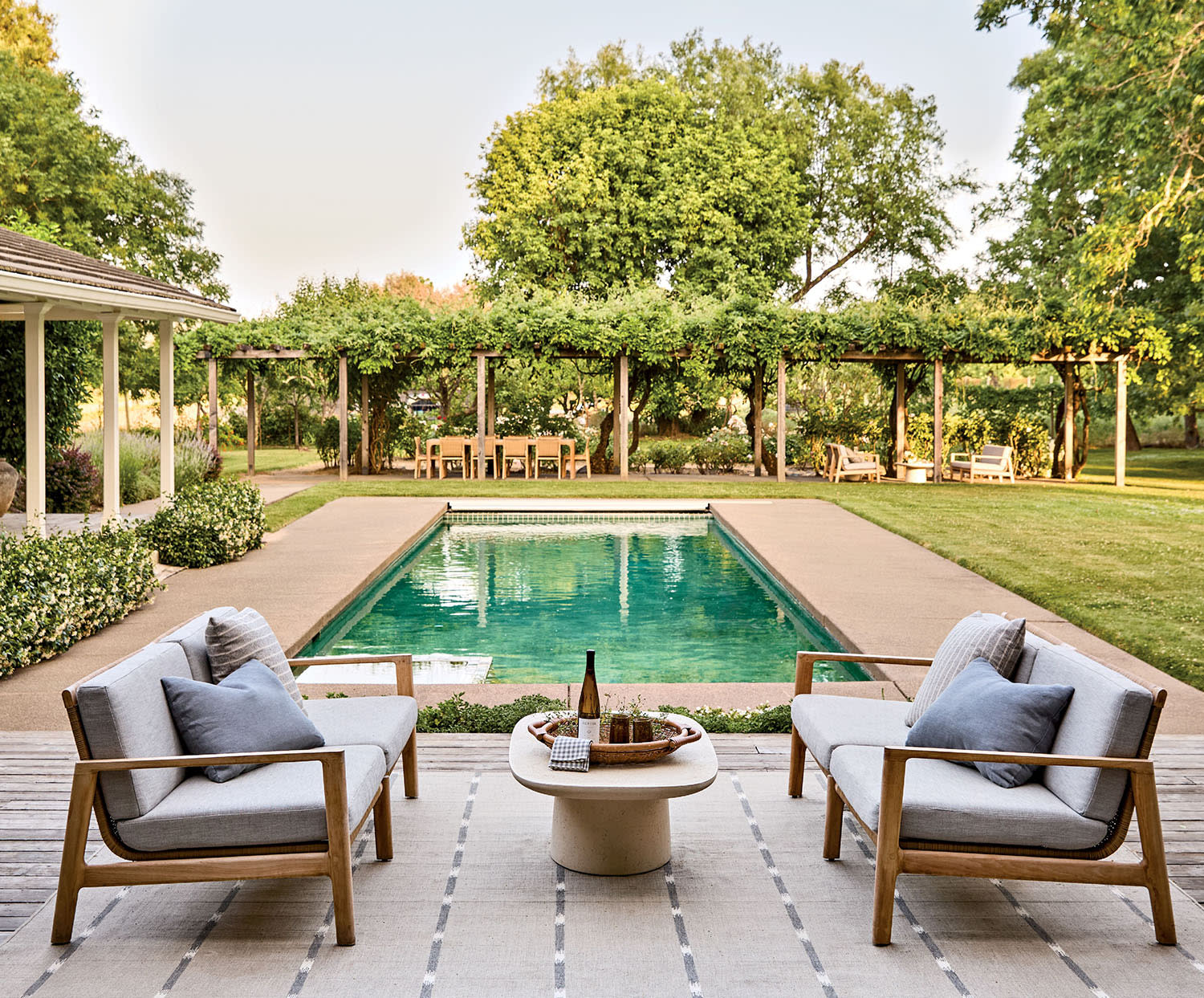 Charming backyard with a pool flanked by loungers and a wooden deck. Lush greenery and pergola create a tranquil, inviting atmosphere.