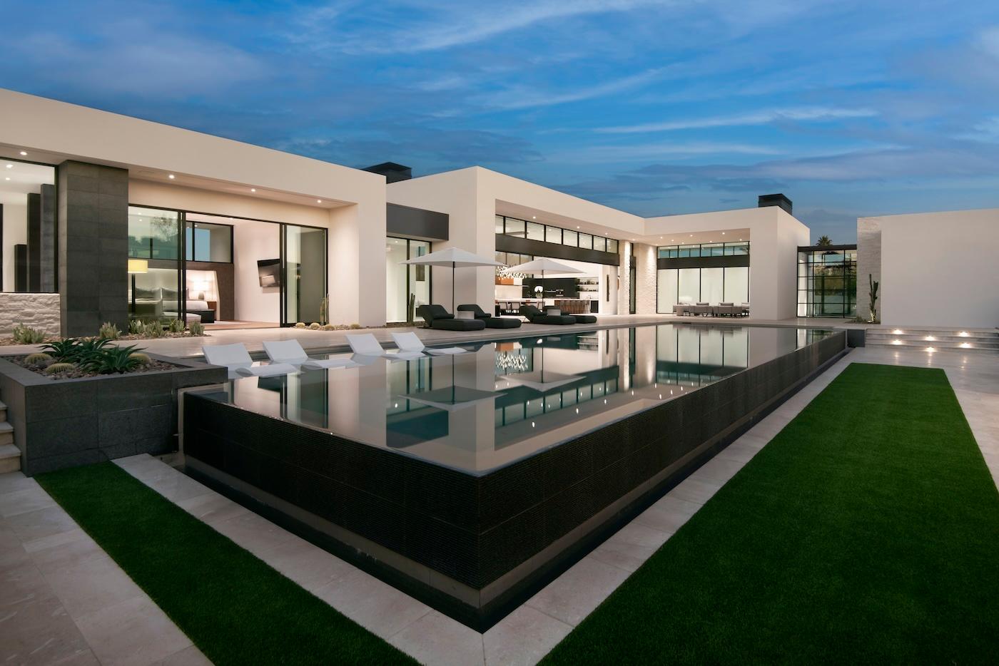 Modern luxury home exterior at dusk with sleek white and gray architecture. Features a large infinity pool, poolside loungers, and glowing interior lights.
