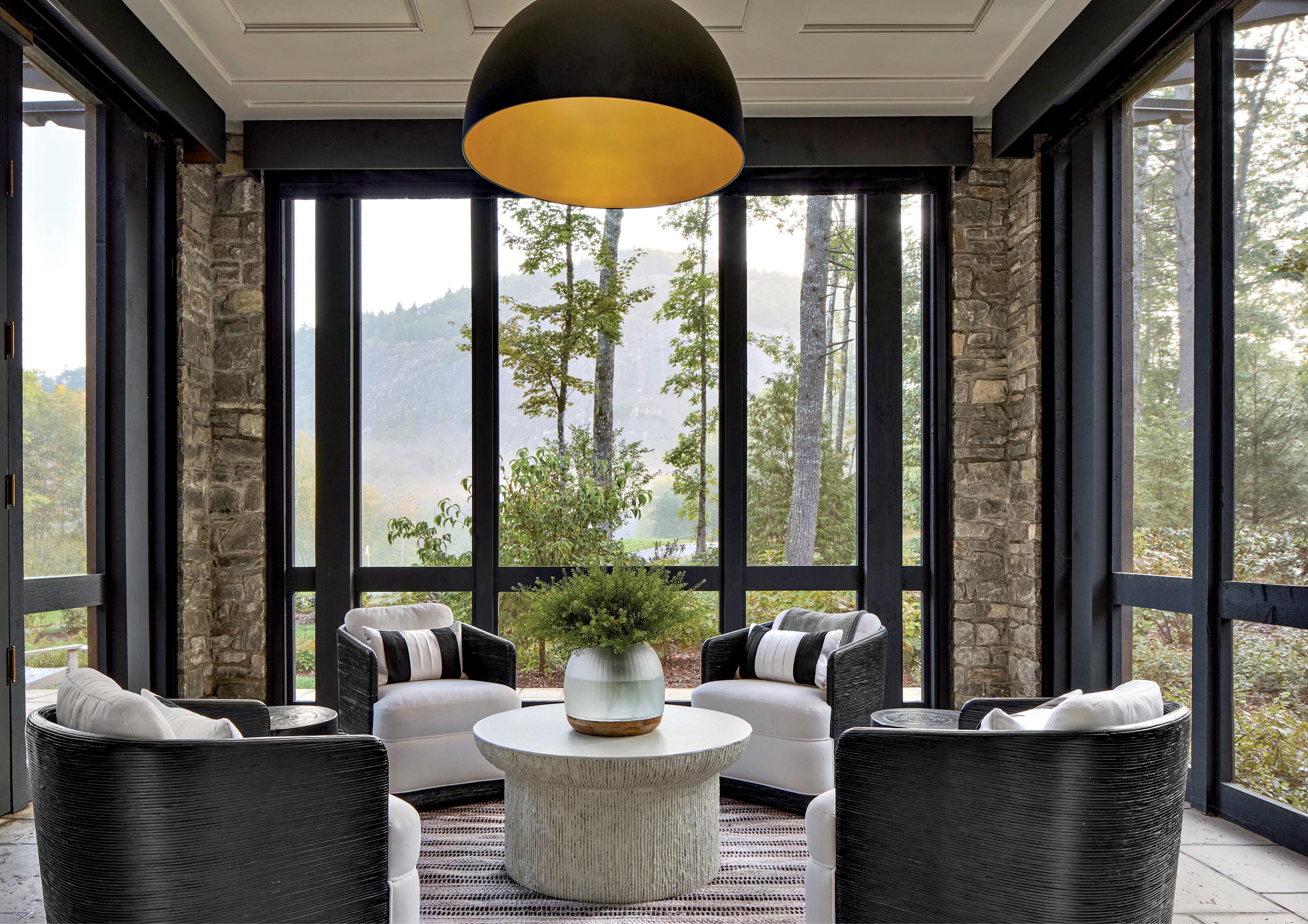 Cozy sunroom with large windows, stone walls, and four black wicker chairs around a round table with a plant. Overhead, a large black lamp emits warm light.