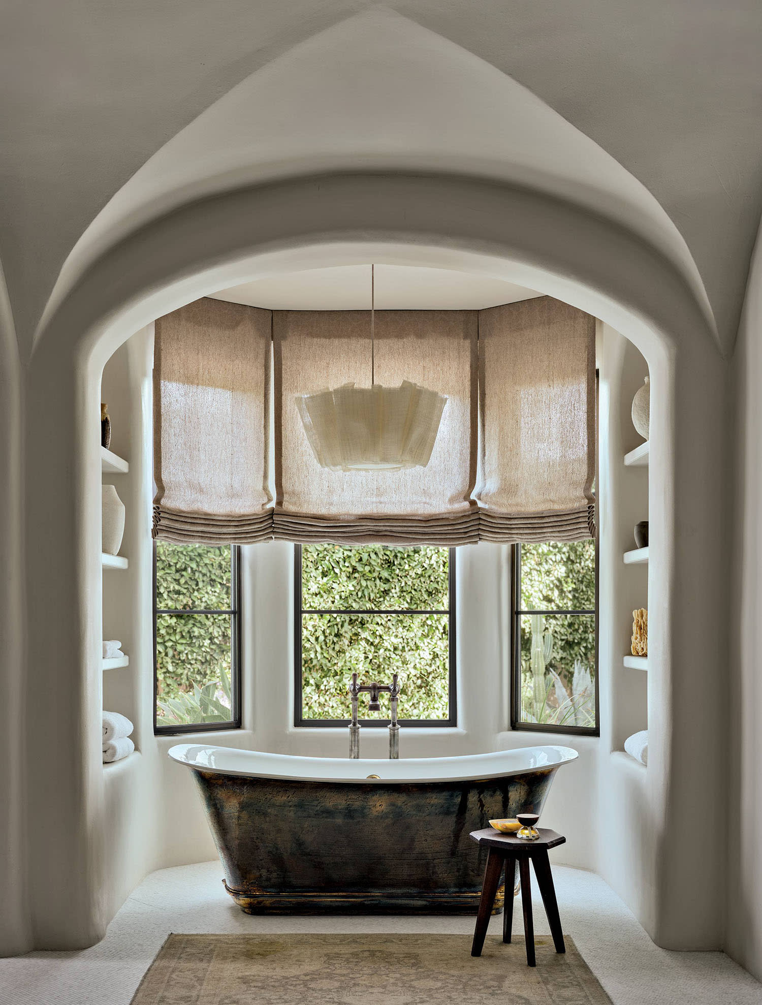 A bathtub sits near a bay window, with shelves on either side and a chandelier hanging above.