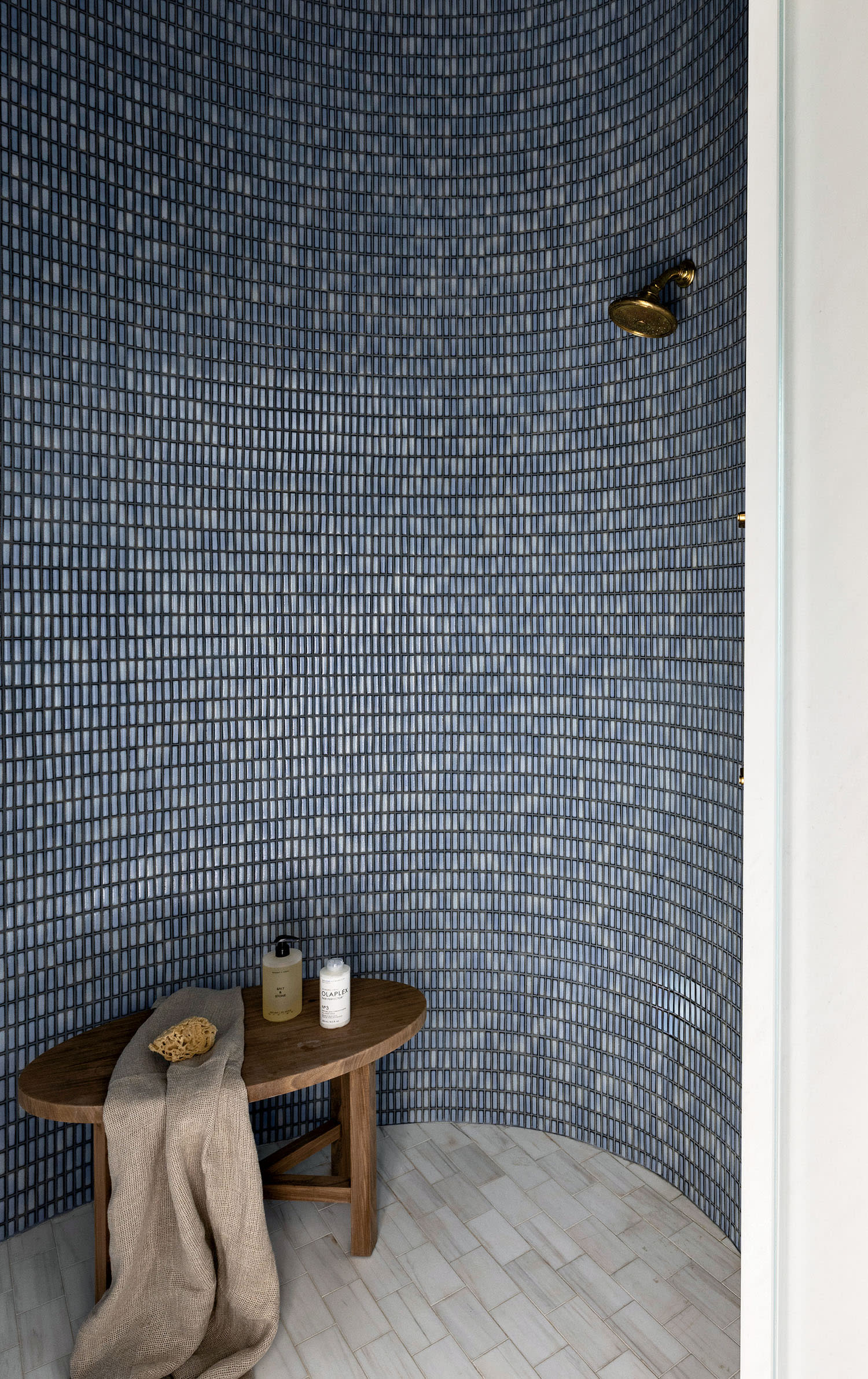 Minimalist bathroom featuring a curved shower with blue mosaic tiles, a brass showerhead, wooden stool with bath items, and a white robe hanging nearby.