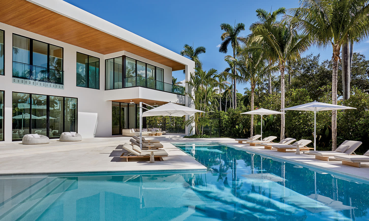 Modern luxury home with large glass windows, surrounded by palm trees, features a serene pool with lounge chairs and umbrellas on a sunny day.