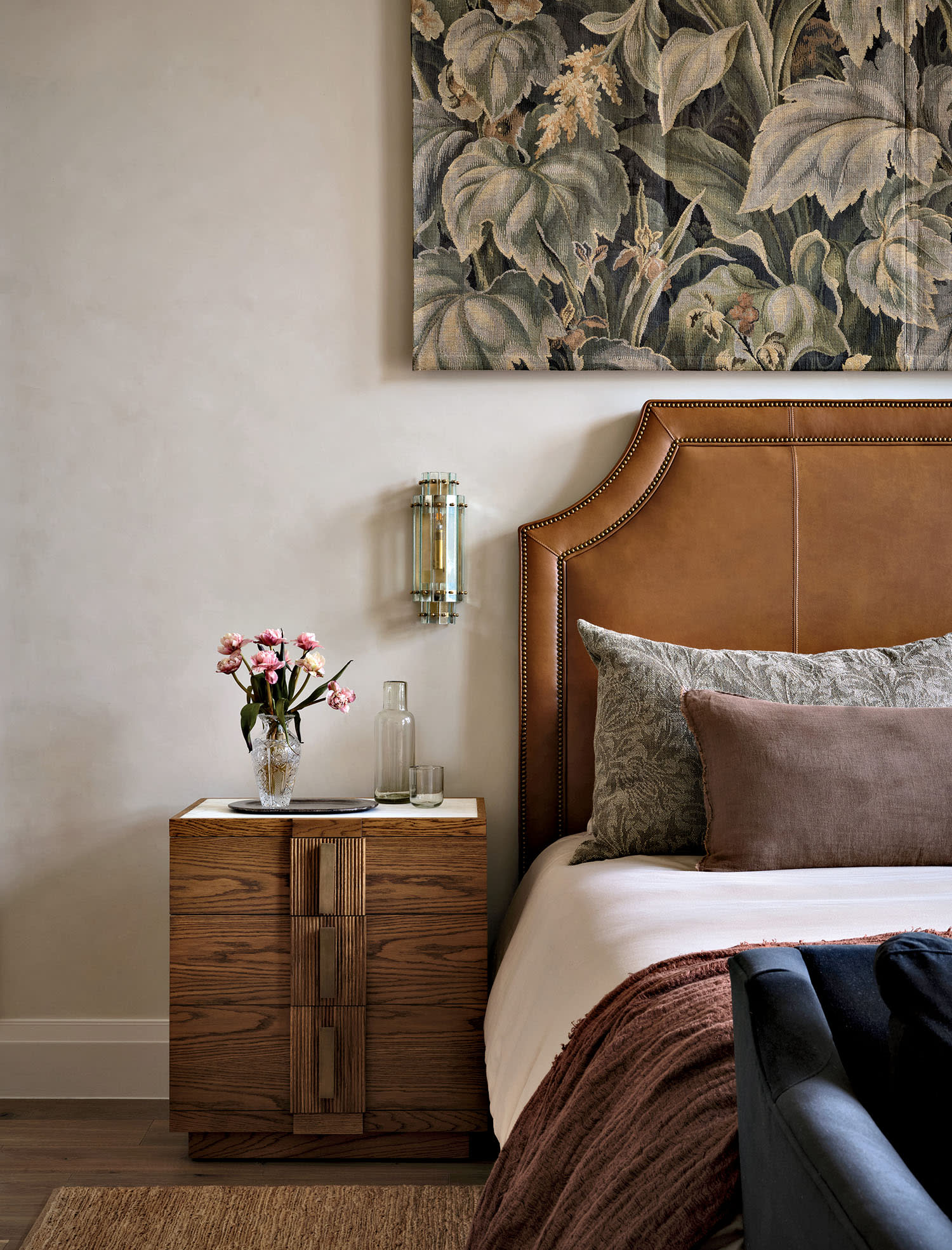 Stylish bedroom with a leather headboard, floral art piece, and a wooden nightstand. Pink flowers in a vase add elegance beside neutral pillows.