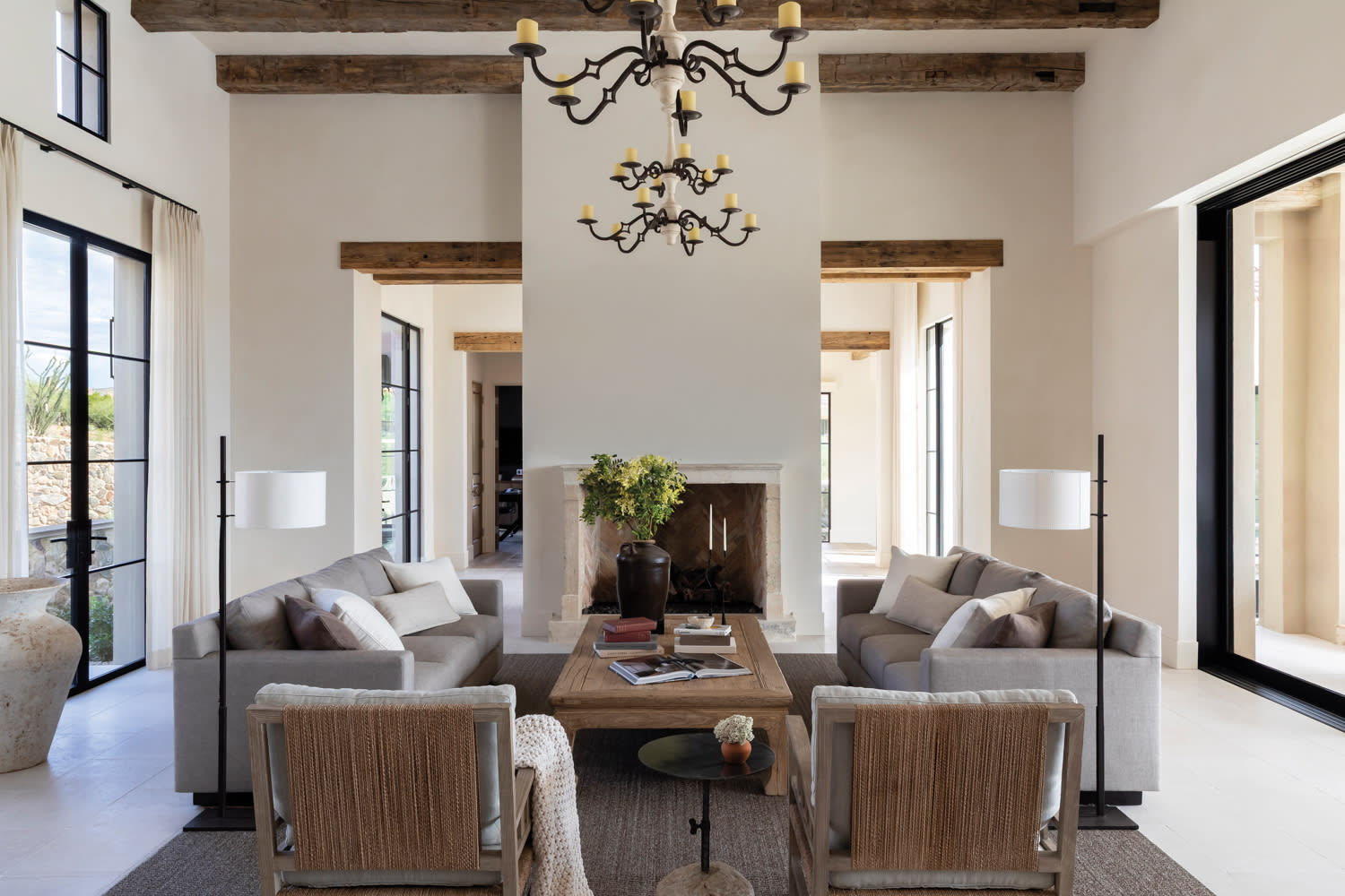 Spacious living room with high ceilings, exposed wooden beams, and a central chandelier. Features neutral-toned sofas, a wooden coffee table, and a fireplace.
