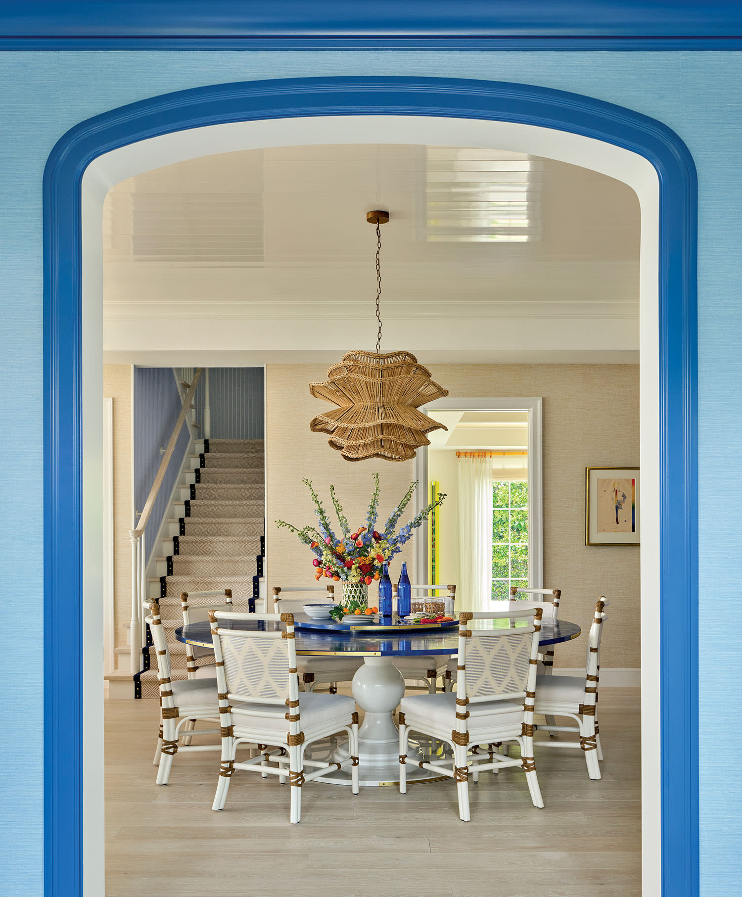 A dining room with blue accents features a round table with white chairs, a floral centerpiece, and a woven pendant light. A staircase and artwork adorn the background.