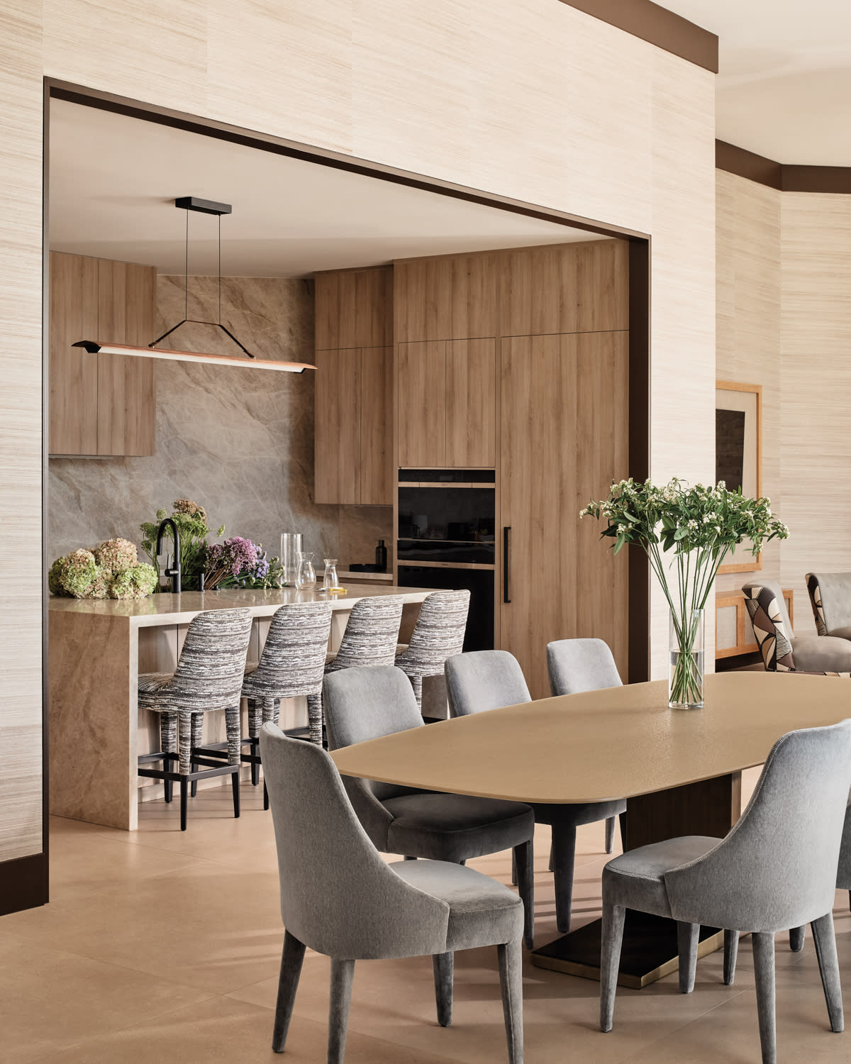 Sleek modern kitchen and dining area with light wood cabinets, stone backsplash, and a long table. Gray chairs, bar stools, and fresh flowers add elegance.