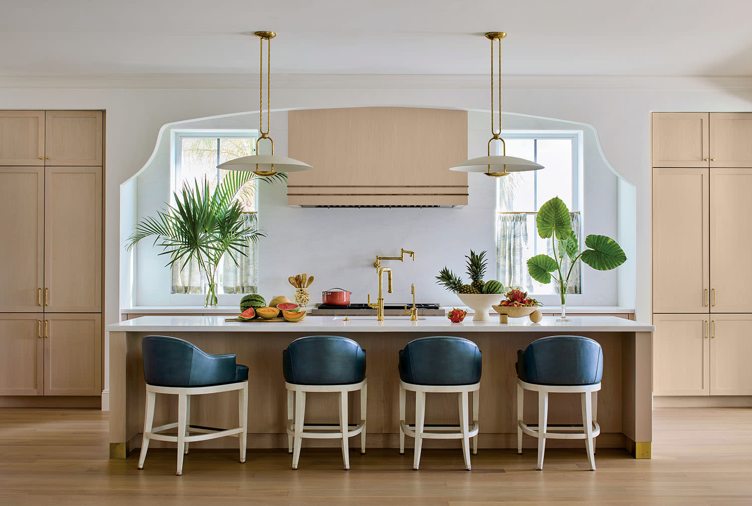 Modern kitchen with a large island featuring blue chairs and gold fixtures. There are two pendant lights, greenery, and a warm, inviting atmosphere.