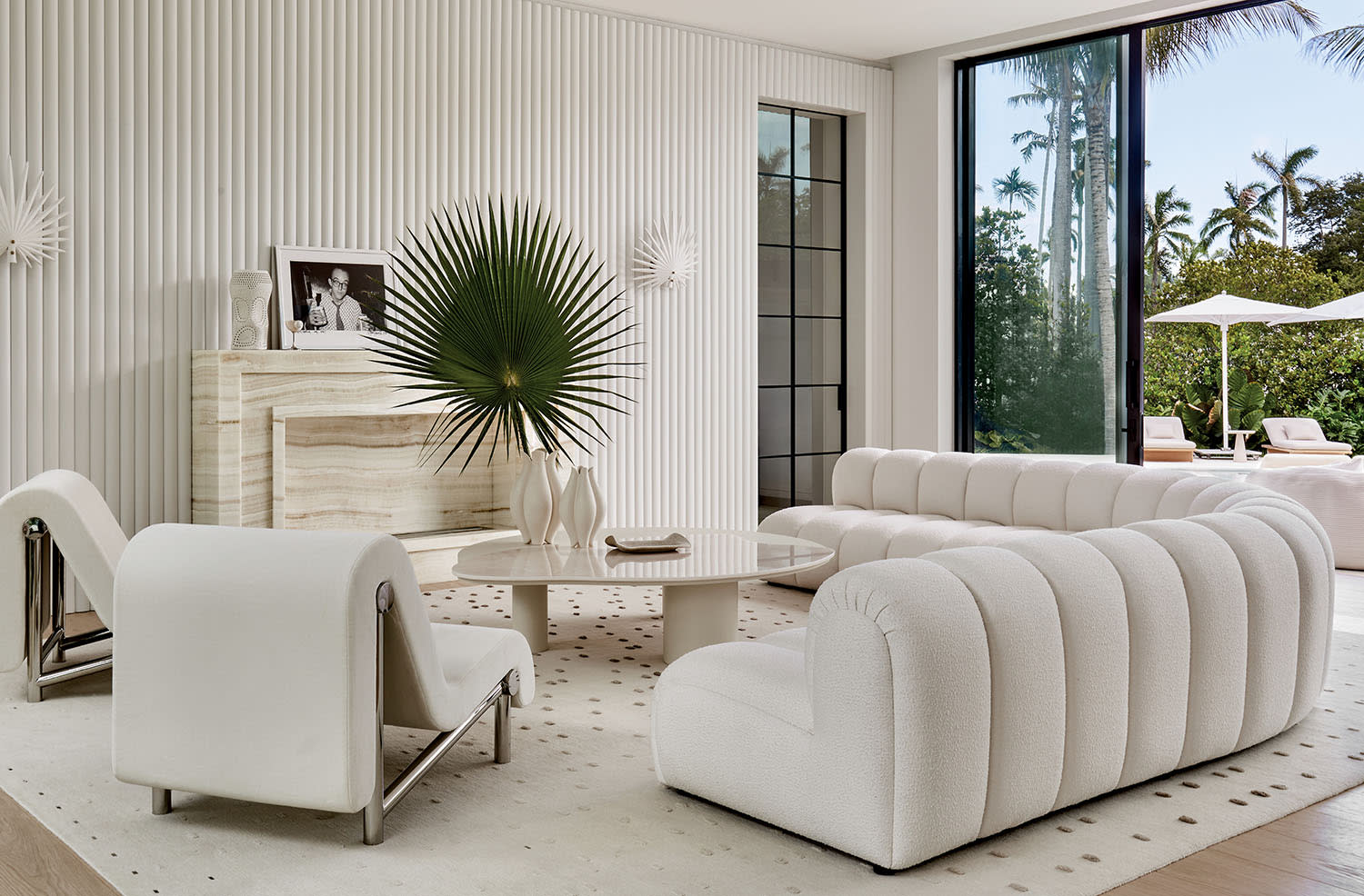 Modern living room with white, plush furniture, a round coffee table, and tropical decor. Large windows reveal a lush, sunny outdoor space.