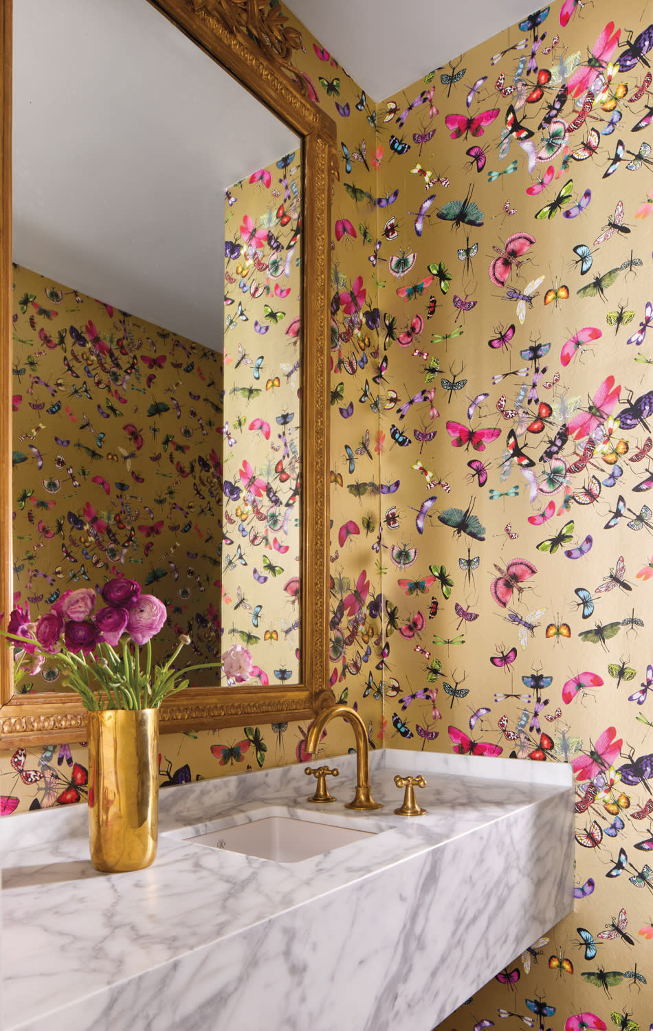 Luxurious bathroom with butterfly-themed wallpaper, a marble sink, golden fixtures, a large ornate mirror, and a gold vase with purple flowers.