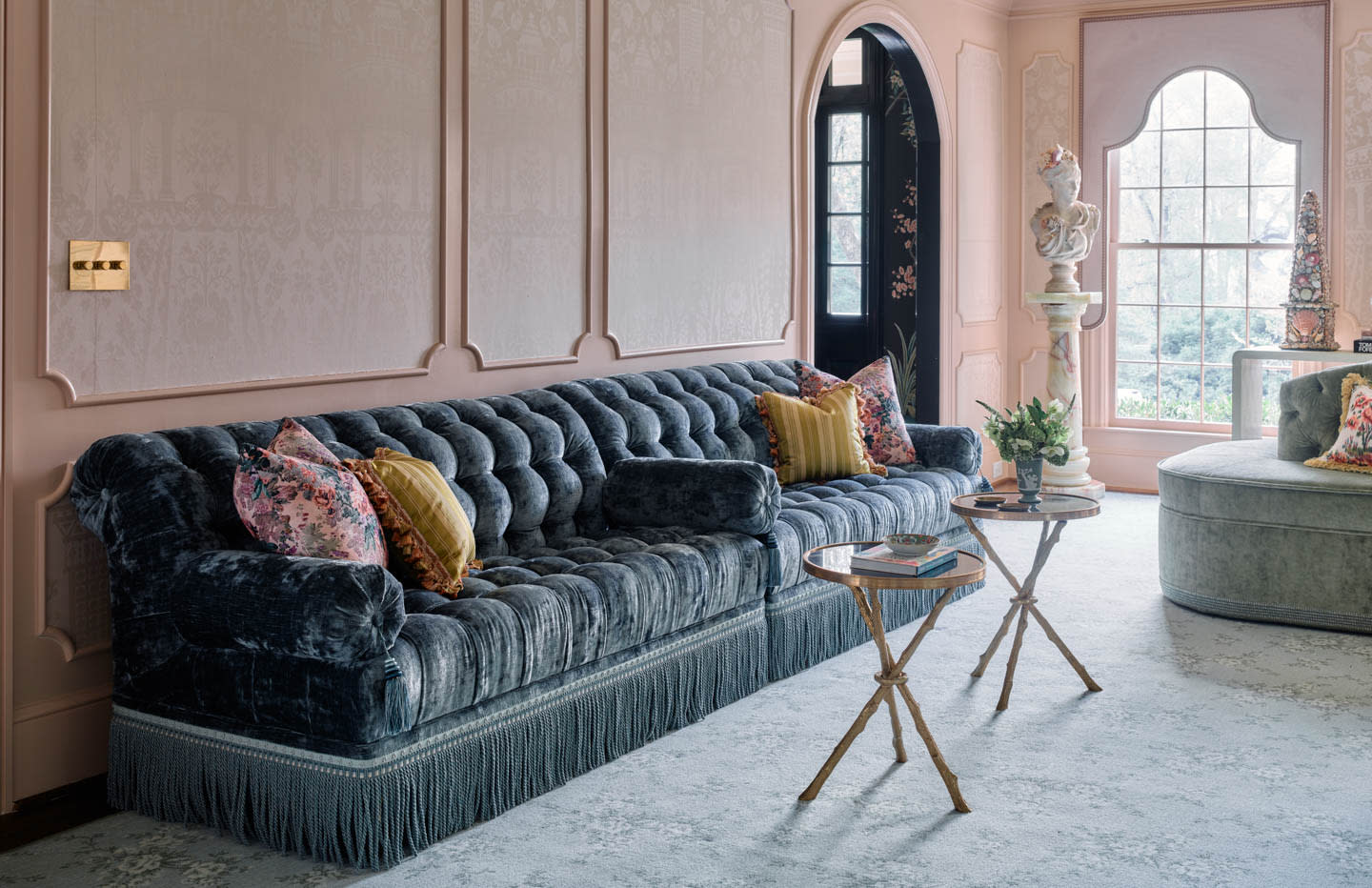 Luxury living room with a plush, dark velvet sofa accented by patterned pillows. Soft pink walls, ornate moldings, and a large window add elegance.
