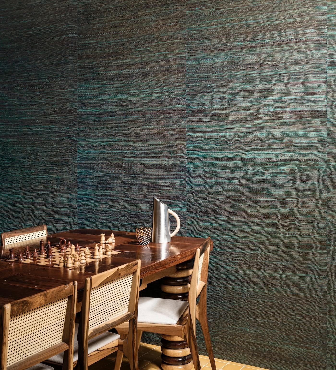 A wooden table with a chessboard set, surrounded by chairs, is beside a textured turquoise and brown wall. A metal pitcher and decorative sphere add modernity.