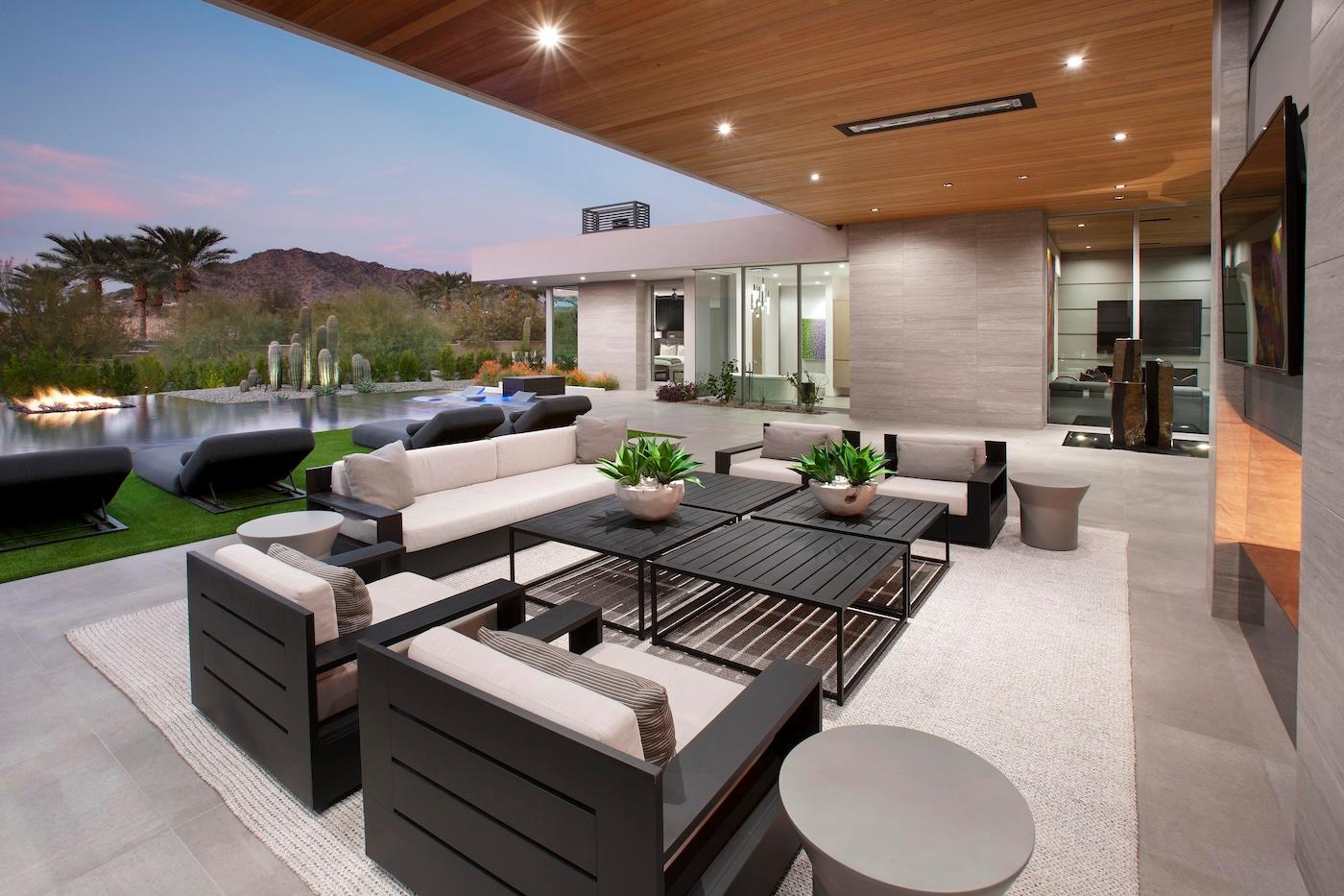 Modern outdoor patio with cushioned seating and black coffee tables, under a wooden ceiling. Nearby, a sleek pool and desert landscape at sunset.