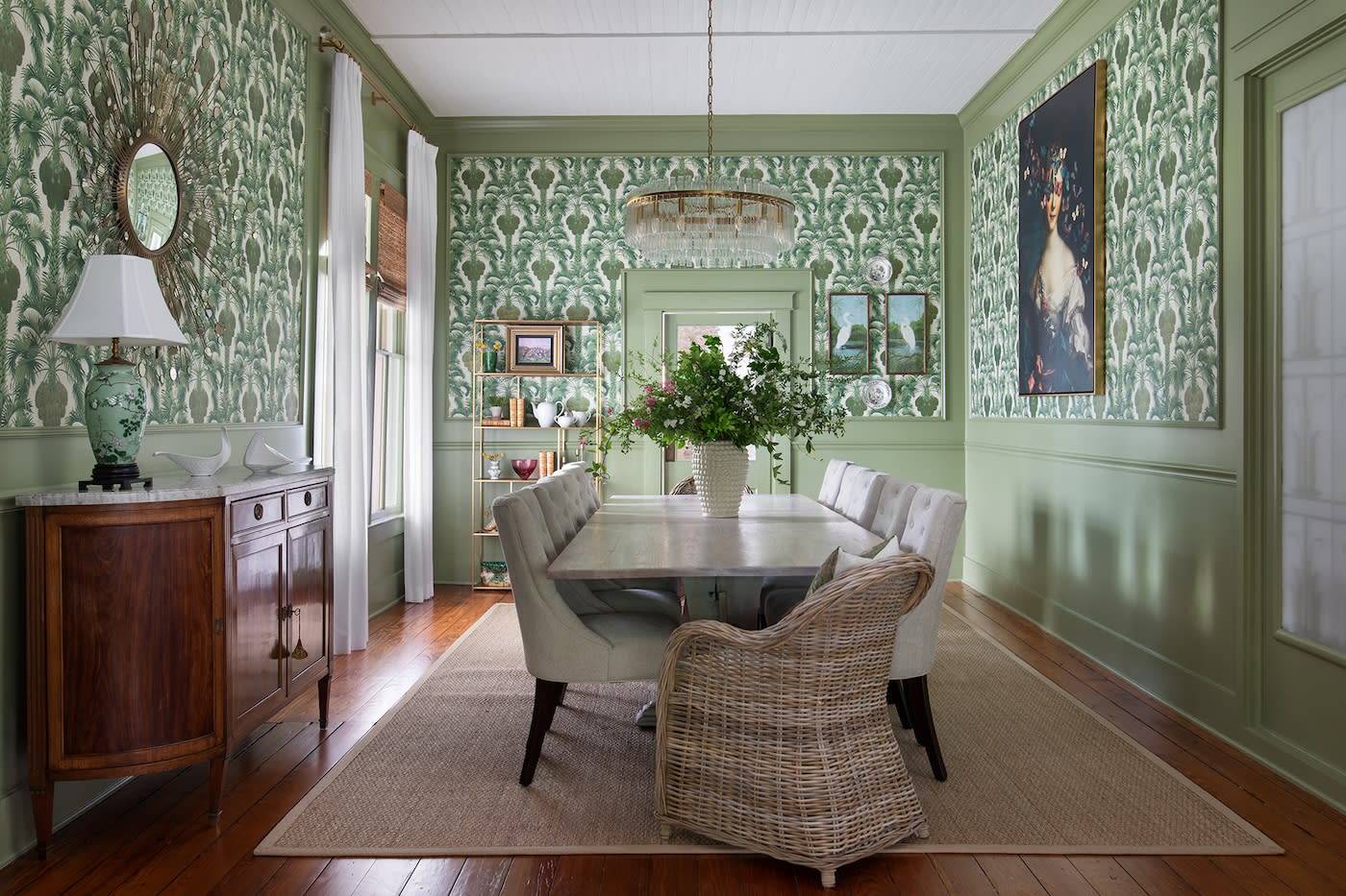 Elegant dining room with green patterned wallpaper, wooden floor, and a large chandelier. A white table surrounded by upholstered chairs and a wicker chair.