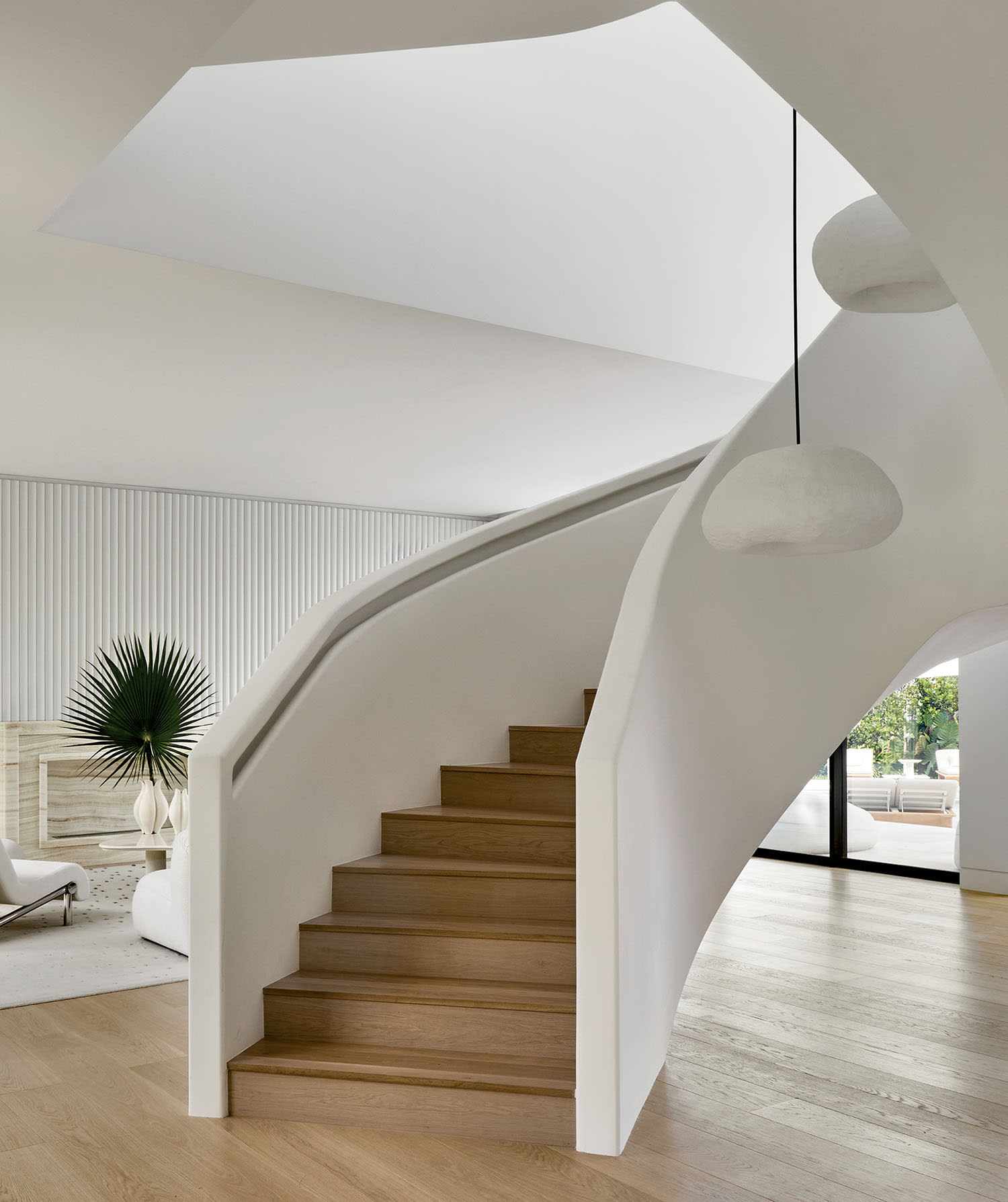 Curved wooden staircase with white walls, modern pendant lights, and a minimalistic living room featuring a white armchair and green plant accents.