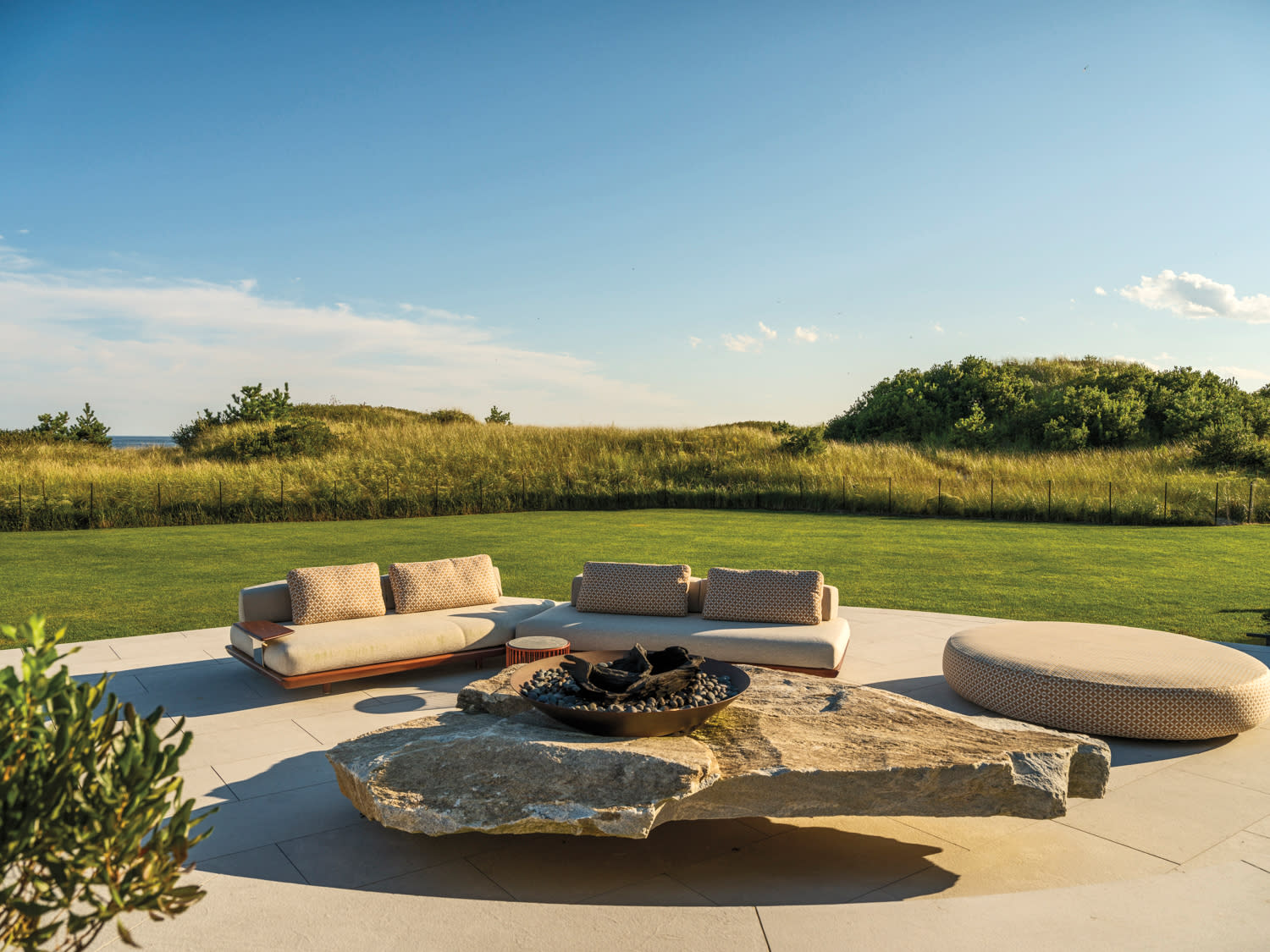 Outdoor patio with modern seating and a stone fire pit, set against a backdrop of lush grass and a clear blue sky. A peaceful and inviting atmosphere.