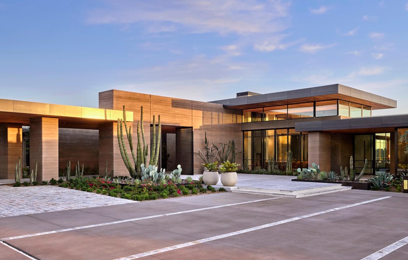 Modern desert home with flat roof and large glass windows, surrounded by cacti and succulents. The setting sun casts warm light, creating a serene atmosphere.