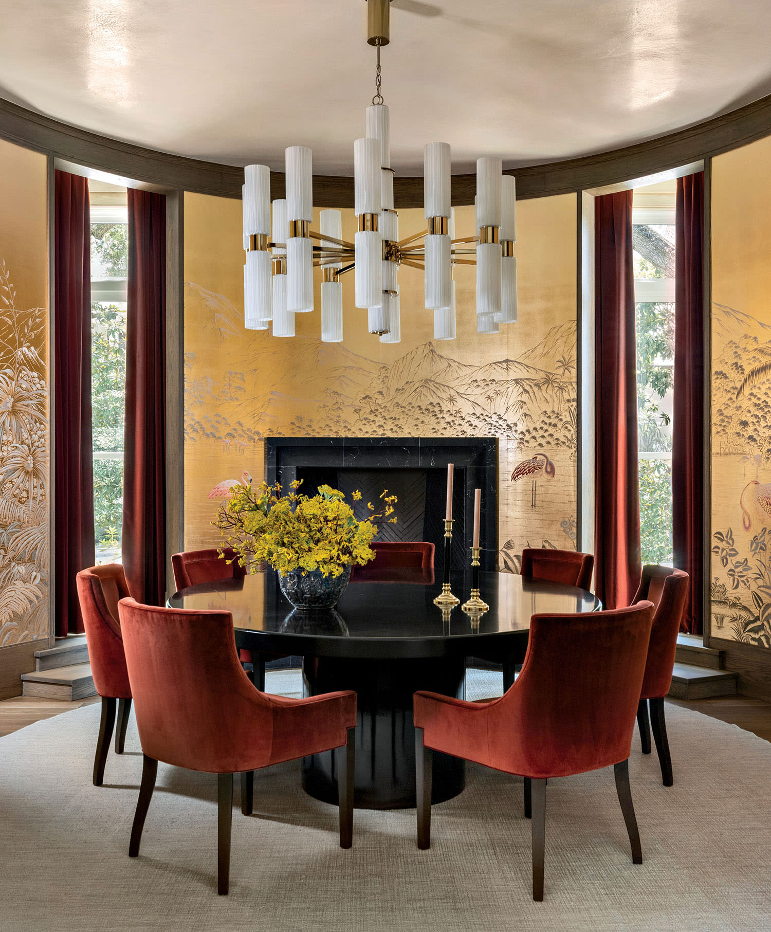Elegant dining room with a round black table, red velvet chairs, and a white chandelier. Gold-toned Asian-inspired mural and yellow flowers add warmth.