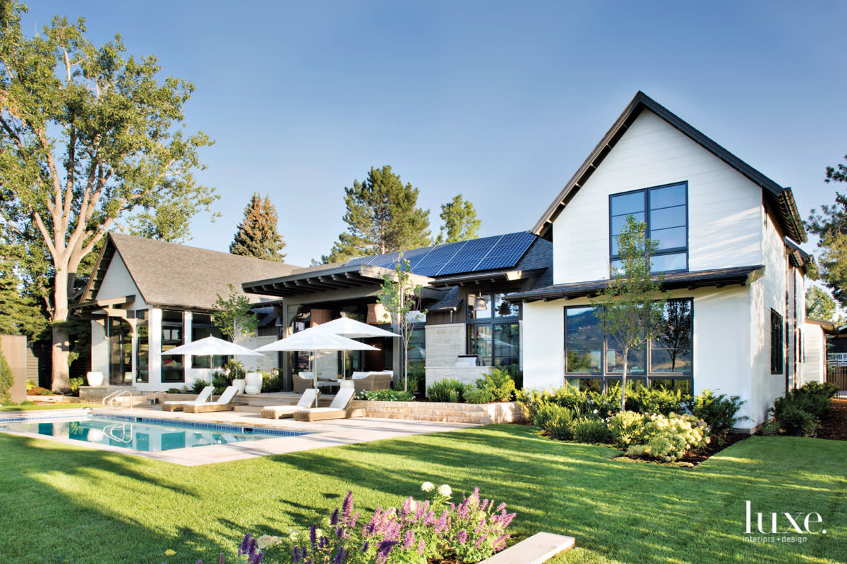 The exterior of a large white home with dark accents has large windows. There's also a large patio and pool.