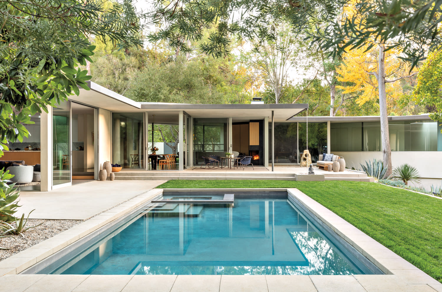 Exterior shot of a rectangular swimming pool that faces a patio with gathering areas and a wing of the house with open window walls