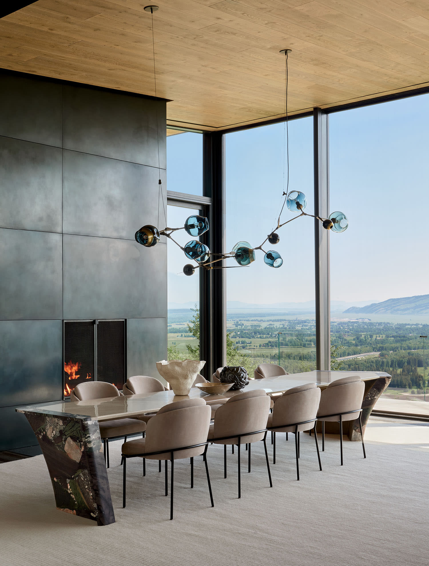 Elegant dining room with a sleek marble table, plush beige chairs, and a modern chandelier with blue glass orbs. Large windows offer a stunning mountain view, while a cozy fire burns in the corner.