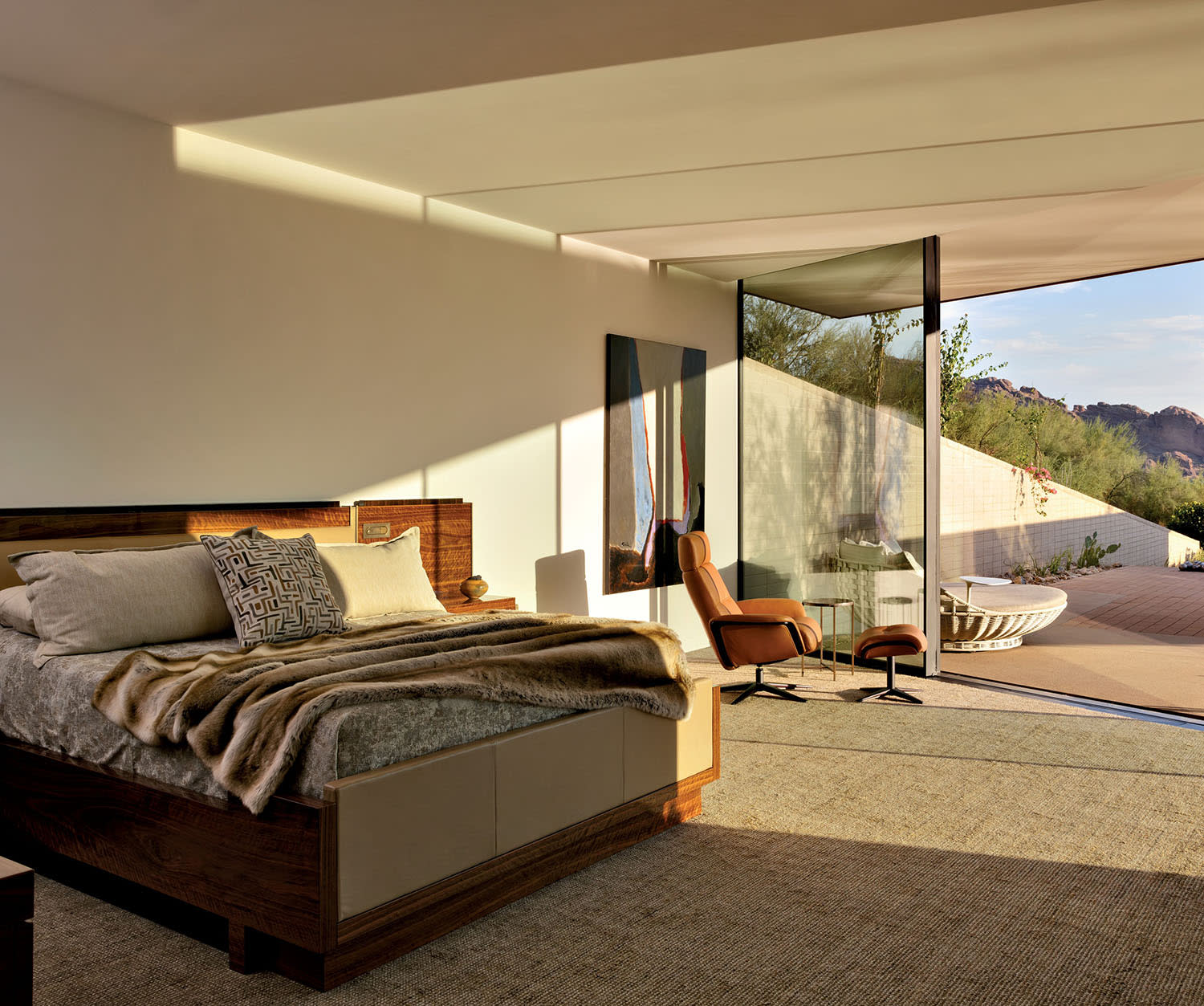 Modern bedroom with glass walls overlooking a sunny desert landscape. Warm tones, a cozy bed with cushions, and an armchair create a serene ambiance.