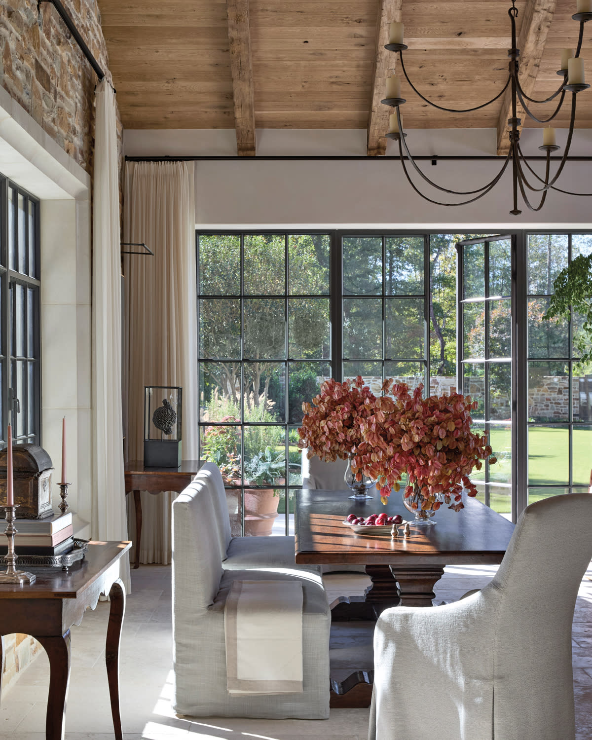 A sunlit dining room with stone walls, tall black-framed windows, and a wooden table adorned with a large bouquet of red leaves. Cozy and elegant.