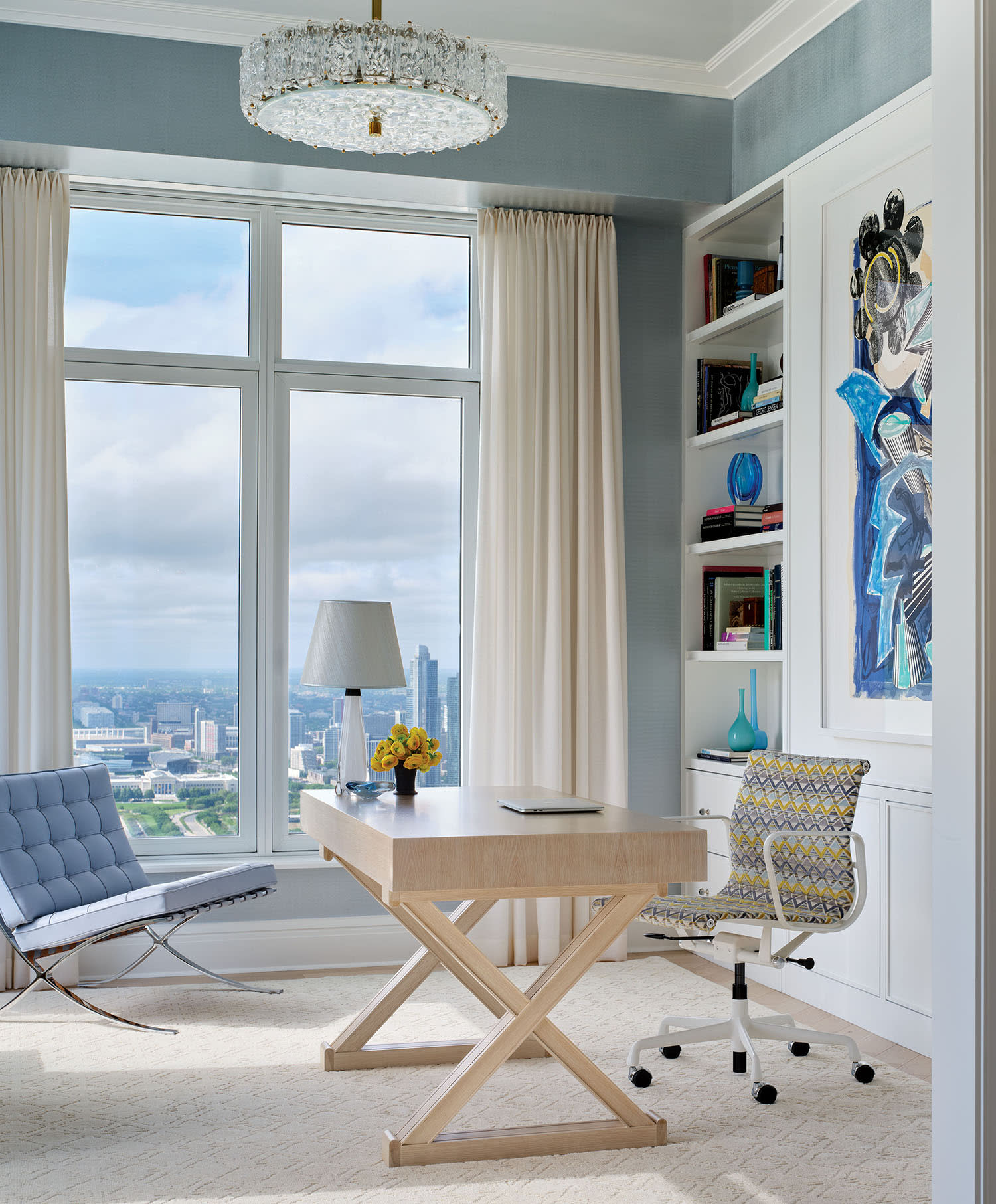 An office space with blue walls and white curtains, with a light-colored wood desk and a blue couch.