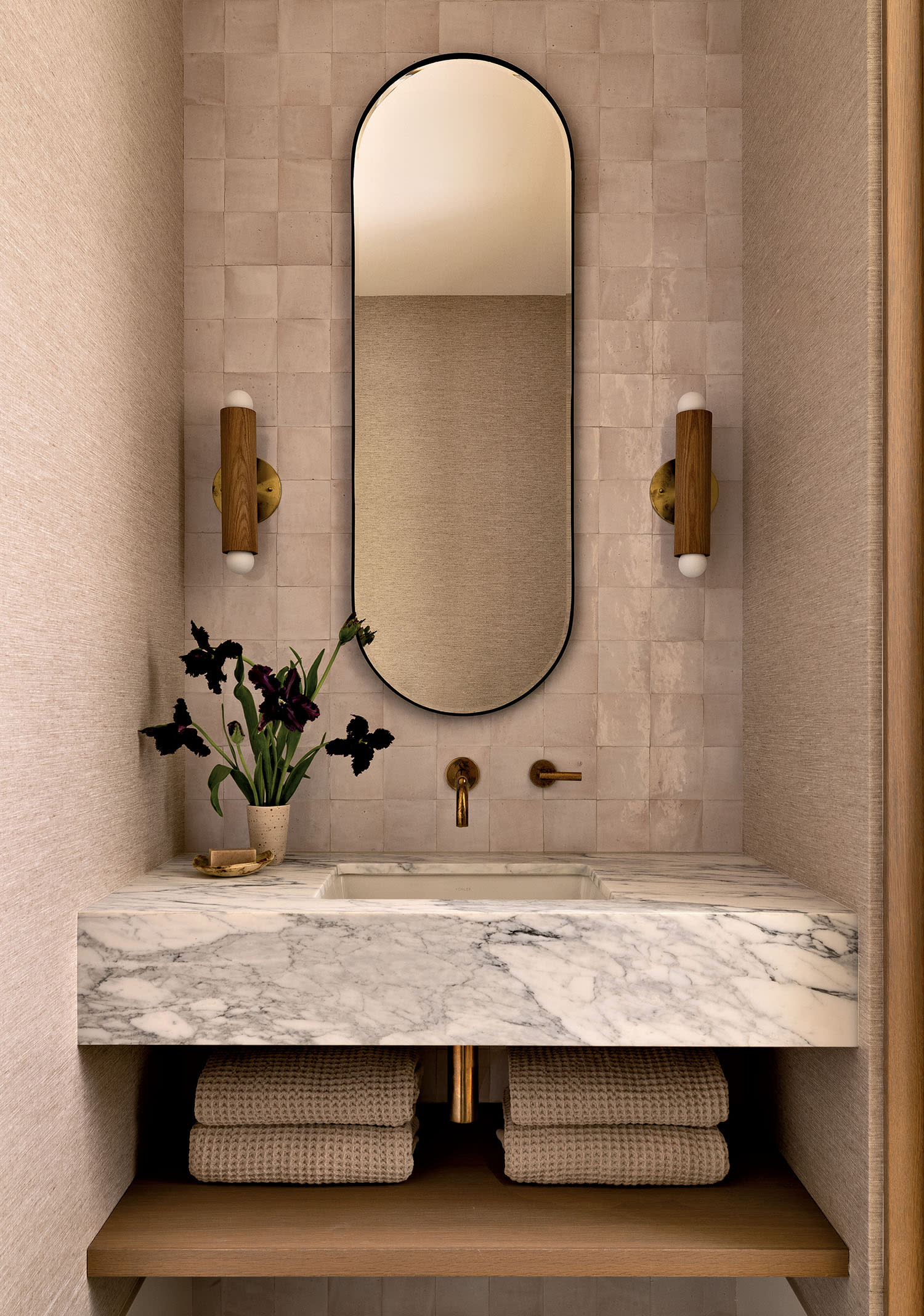 Elegant bathroom with a marble sink, brass fixtures, and oval mirror. Dark flowers in a vase add contrast. Calm, stylish ambiance.