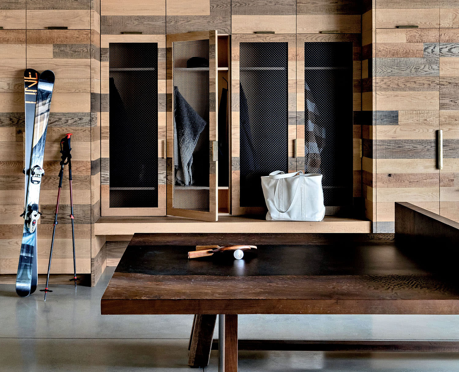Rustic sports-themed room with wooden lockers, ski equipment, and a large wooden table. Warm lighting and a cozy rug enhance a welcoming ambiance.