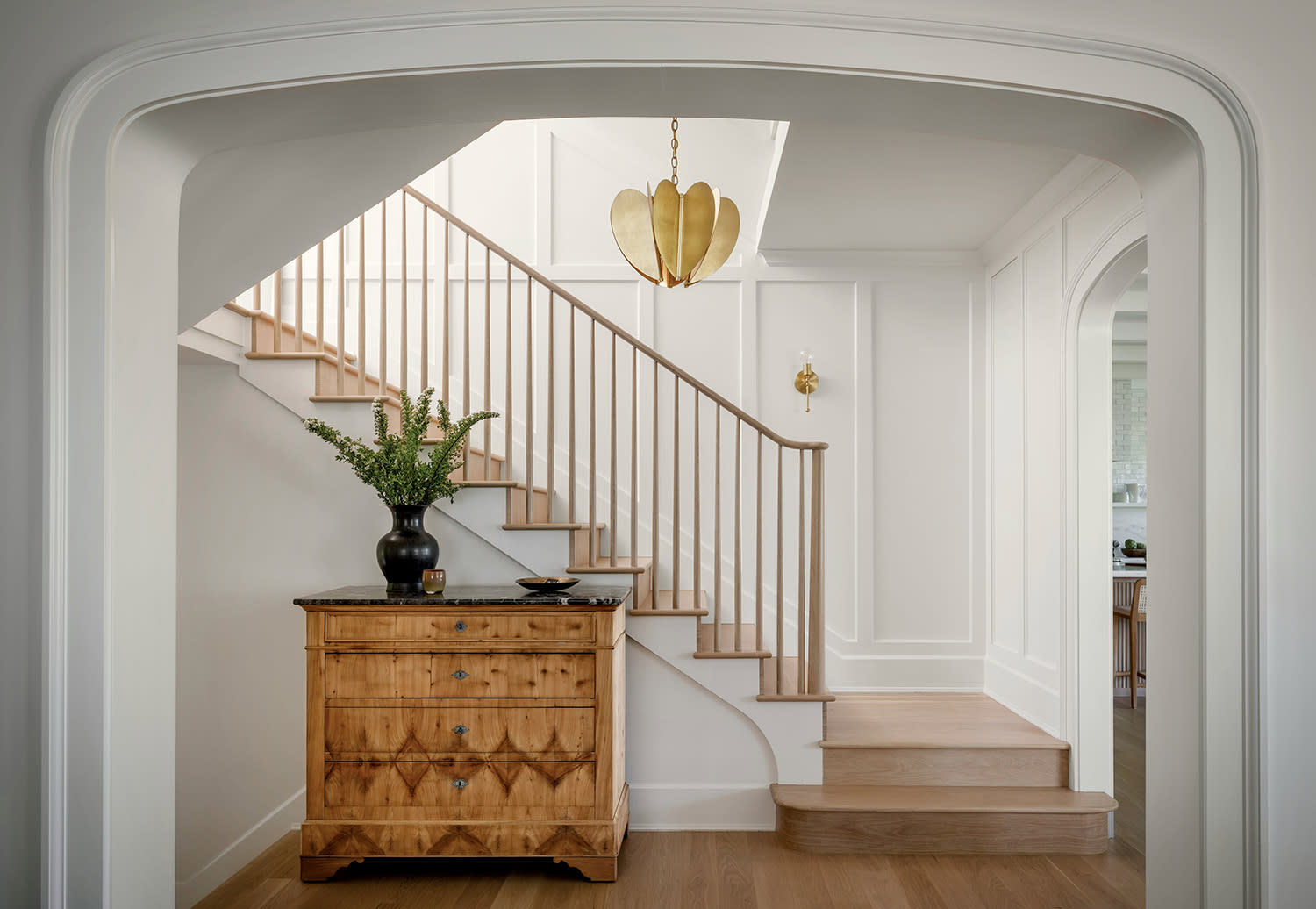Elegant foyer with wooden stairs and a warm-tone chest of drawers. A gold pendant light hangs above, casting a cozy glow, enhancing the tranquil atmosphere.