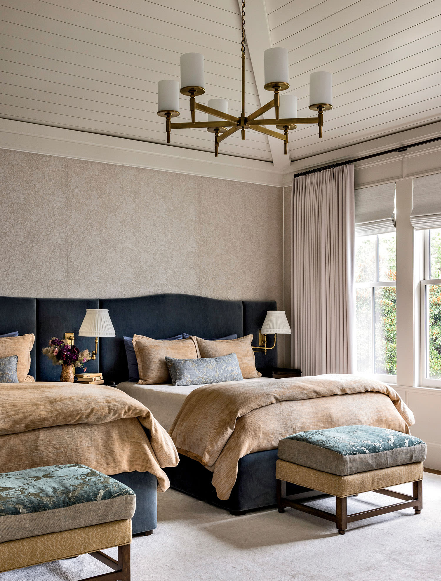 Elegant bedroom with two plush beds featuring beige and blue bedding, flanked by nightstands with lamps. A brass chandelier hangs from the ceiling, and a large window with soft curtains lets in natural light