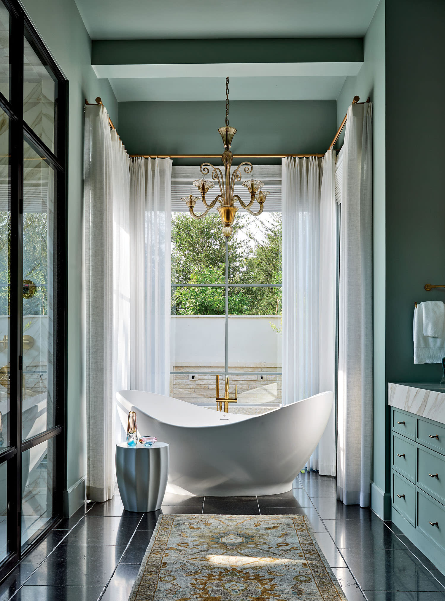 Luxurious bathroom with an elegant white freestanding tub under a gold chandelier. Large window with sheer curtains, black tile floor, and light blue walls.