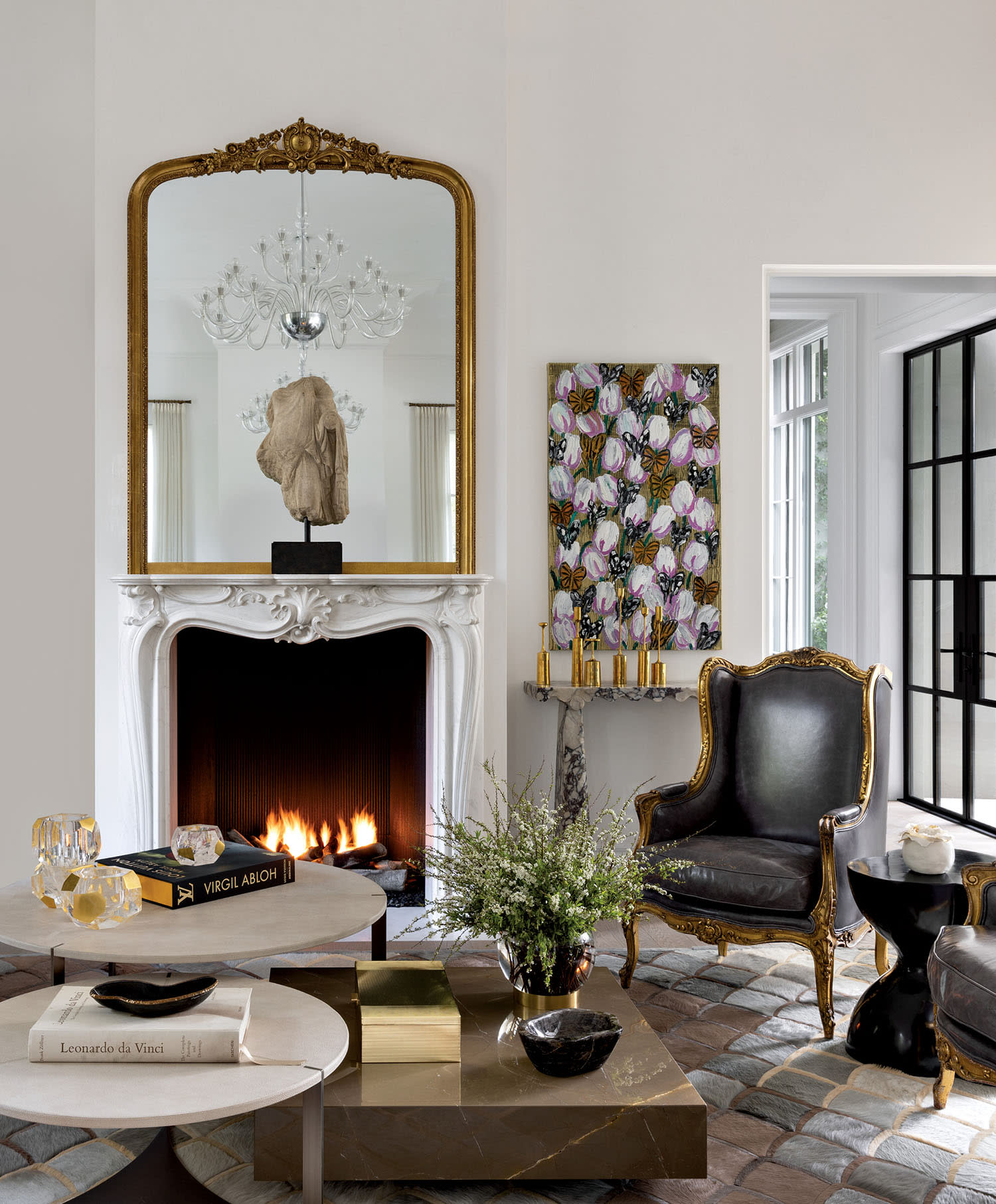 Elegant living room with a lit fireplace beneath an ornate mirror. A black armchair with gold accents, floral artwork, and chic decor convey sophistication.