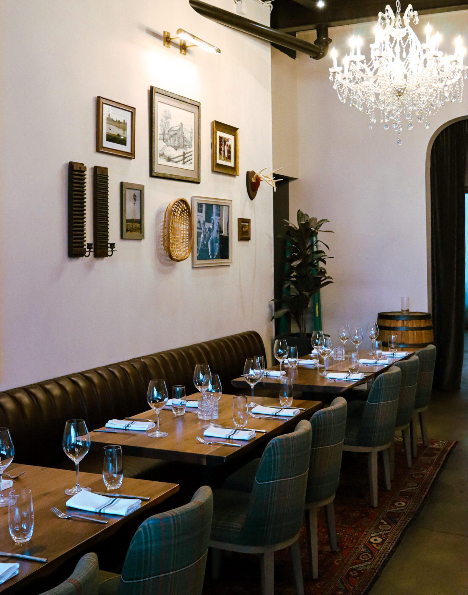 Restaurant dining space with 3 wood-topped tables lined with a banquette on one side and chairs on the other underneath a crystal chandelier. 