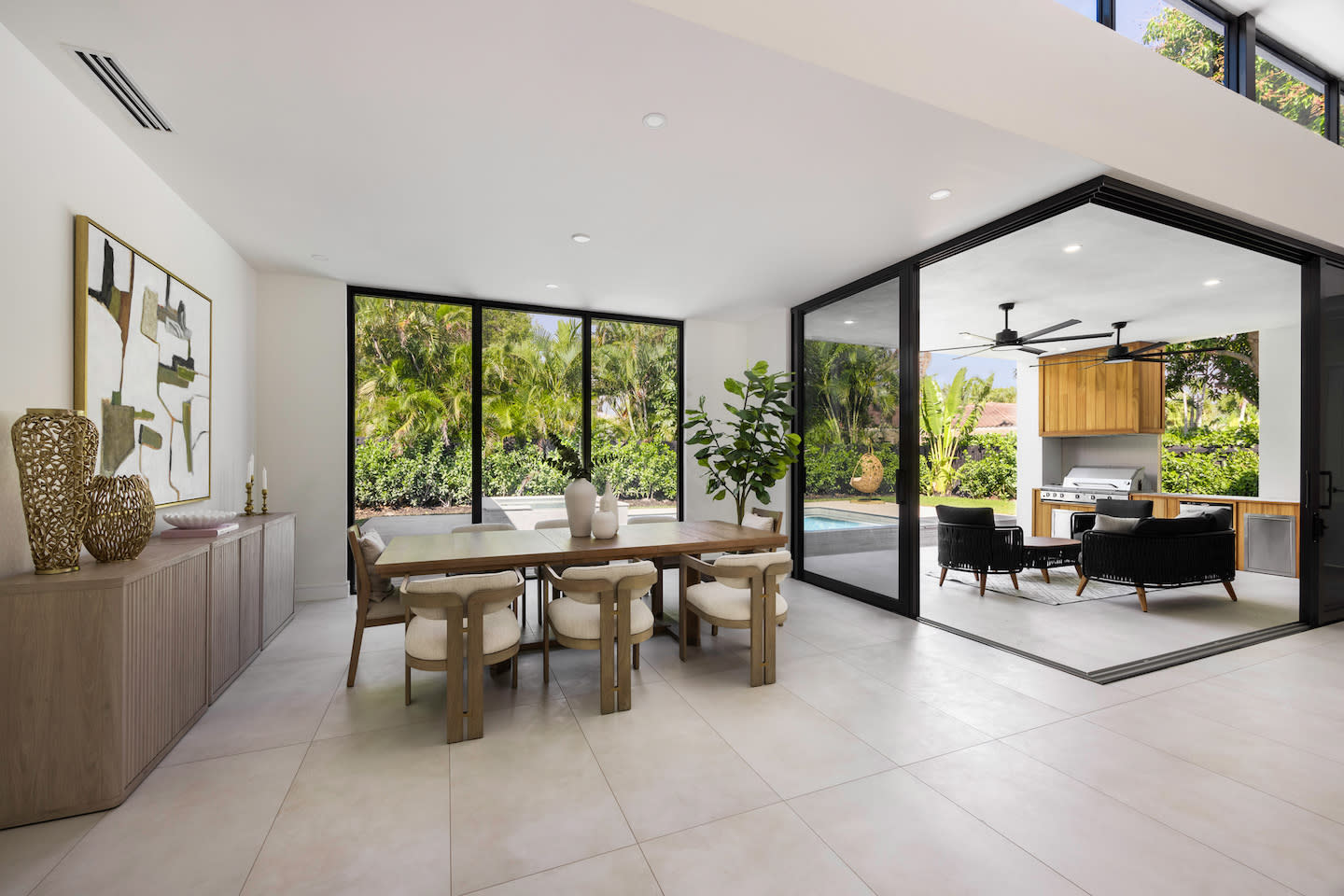Bright indoor dining area with a natural-oak table and pillowy cream chairs, set beside floor-to-ceiling black-framed windows that overlook a palm-ringed pool. Folding glass doors disappear to merge the space with a covered patio featuring a built-in wood-clad grill station, ceiling fans, and woven lounge chairs, creating a seamless indoor-outdoor vibe. Sleek porcelain floors, abstract art, and a low credenza with sculptural vases complete the airy, modern look.