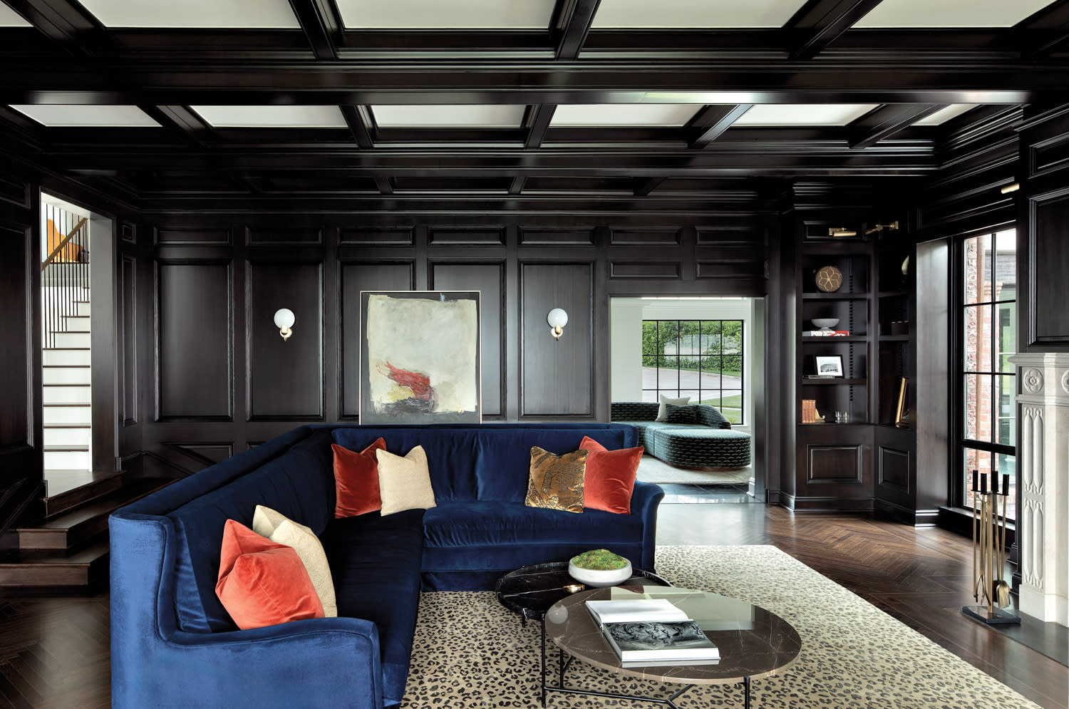 Living room with dark paneling and beams, velvet blue sectional, coffee tables and white sconces next to a painting